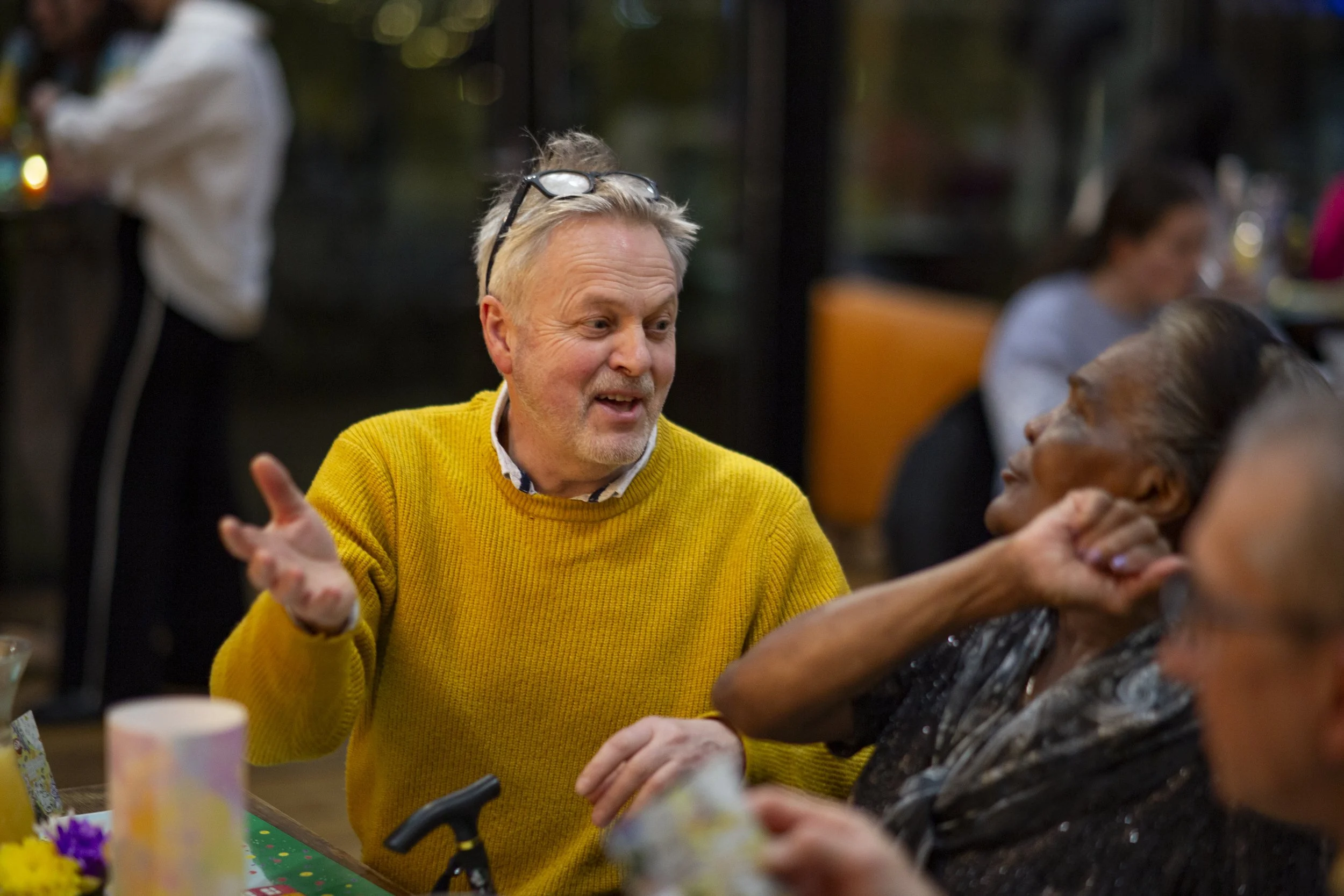 A man in a yellow sweater is talking and gesturing with his hands while a woman with gray hair and dark skin listens. There are other people in the background at what appears to be a social gathering.