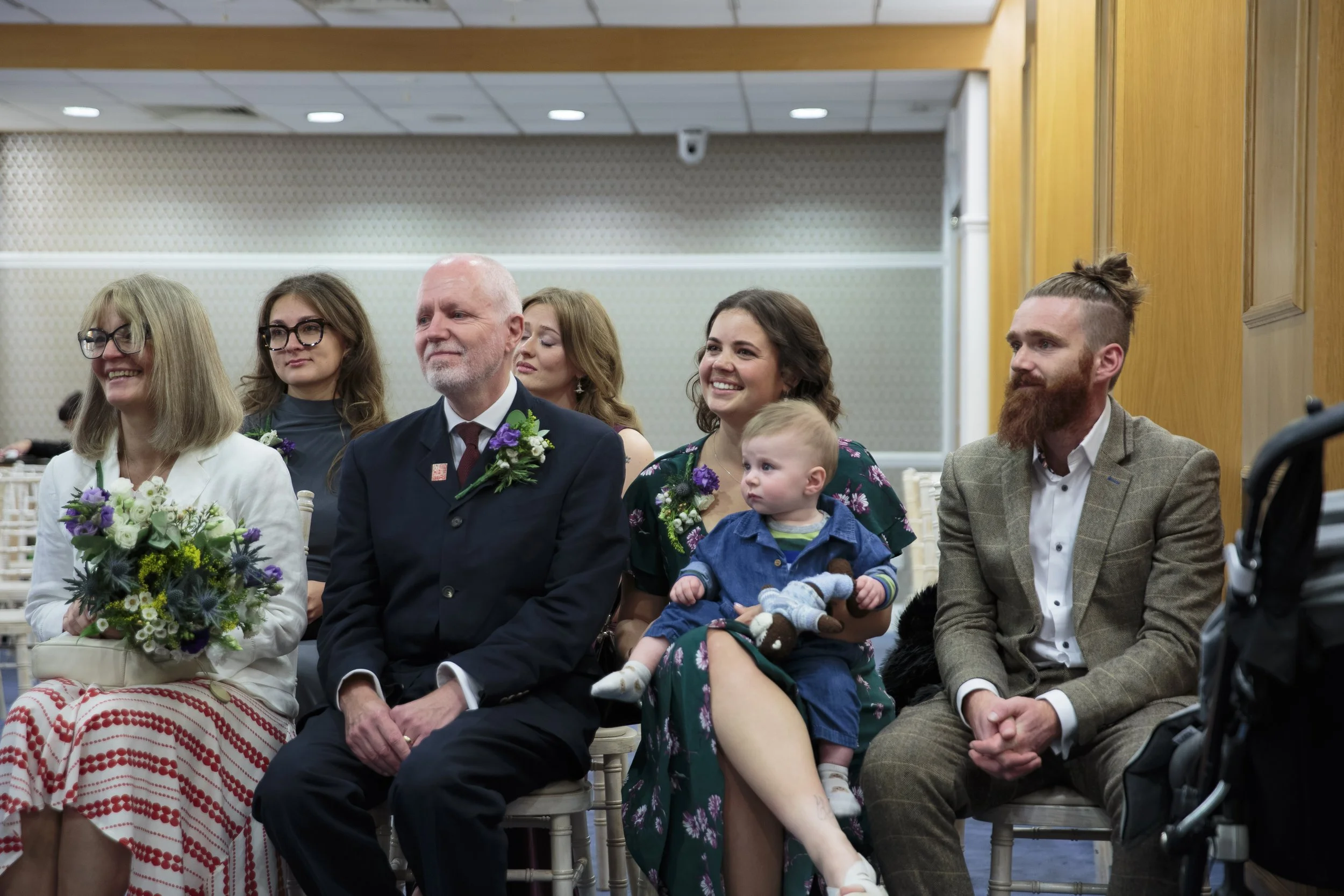 Group of people sitting in a room, dressed in wedding attire, attending a wedding ceremony. Some are smiling, and a woman in the front holds a bouquet of flowers.