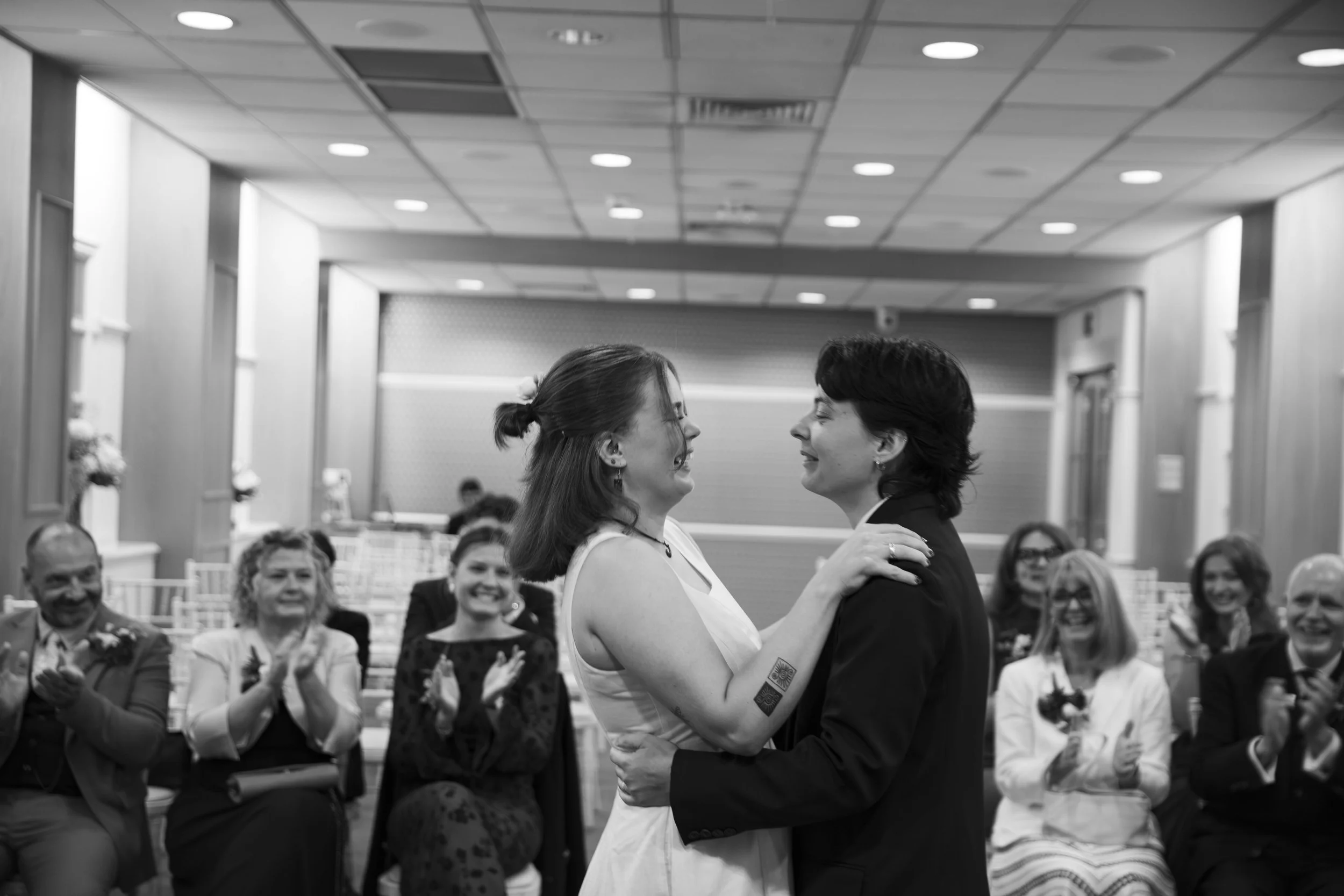 Two women dancing closely at a wedding reception, with guests clapping and smiling in the background.