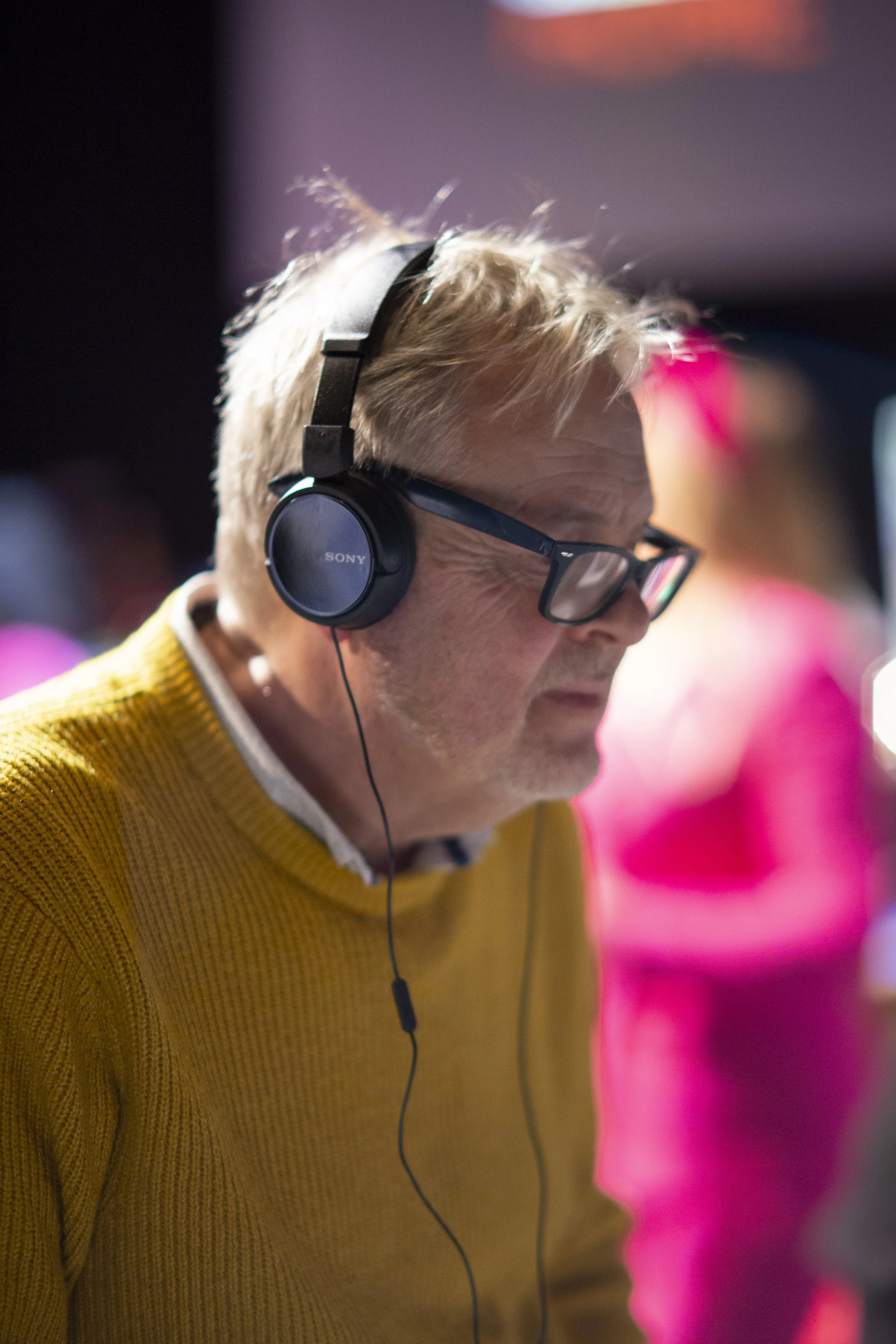 A man with glasses and blonde hair wearing a yellow sweater, listening with headphones.