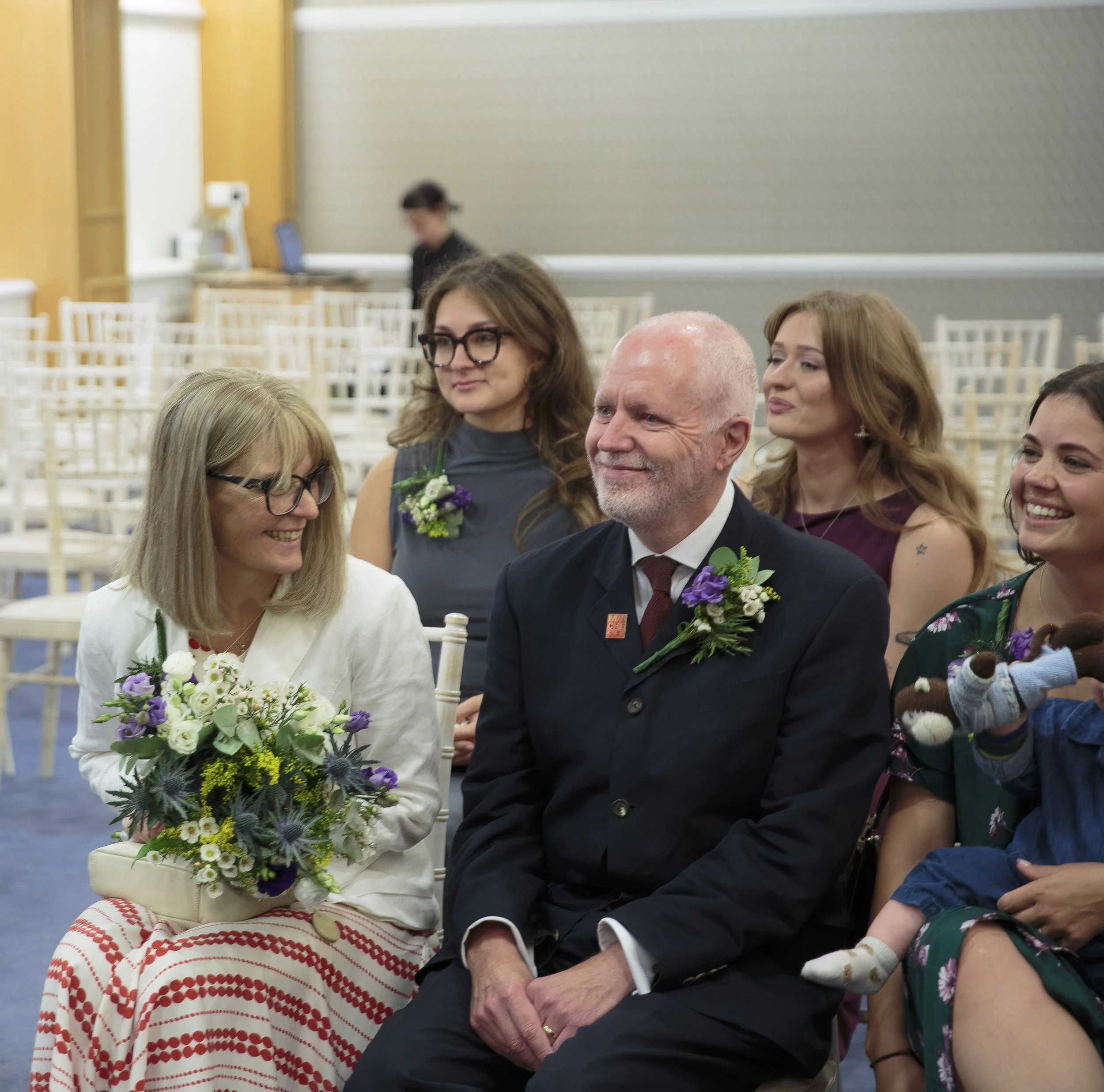 People at a celebration, likely a wedding, sitting together and smiling. The woman in the foreground is holding a bouquet of flowers, and a man in a suit is nearby. Other women are visible around them.