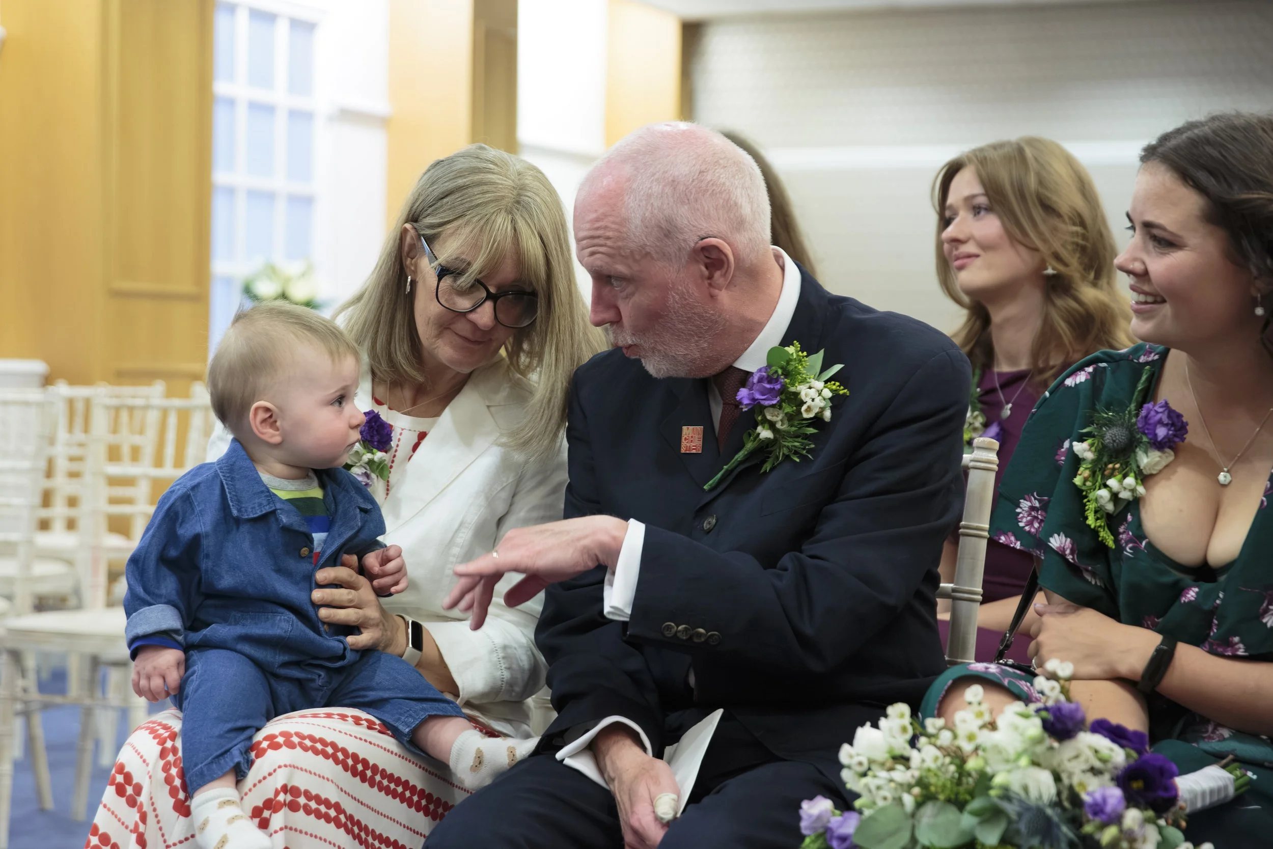 A photo of a wedding ceremony in an indoor location. An elderly man and a young child are sitting together, engaged in a close conversation. The man is wearing a dark suit with flower boutonniere and a pin on his lapel. The woman next to the child is