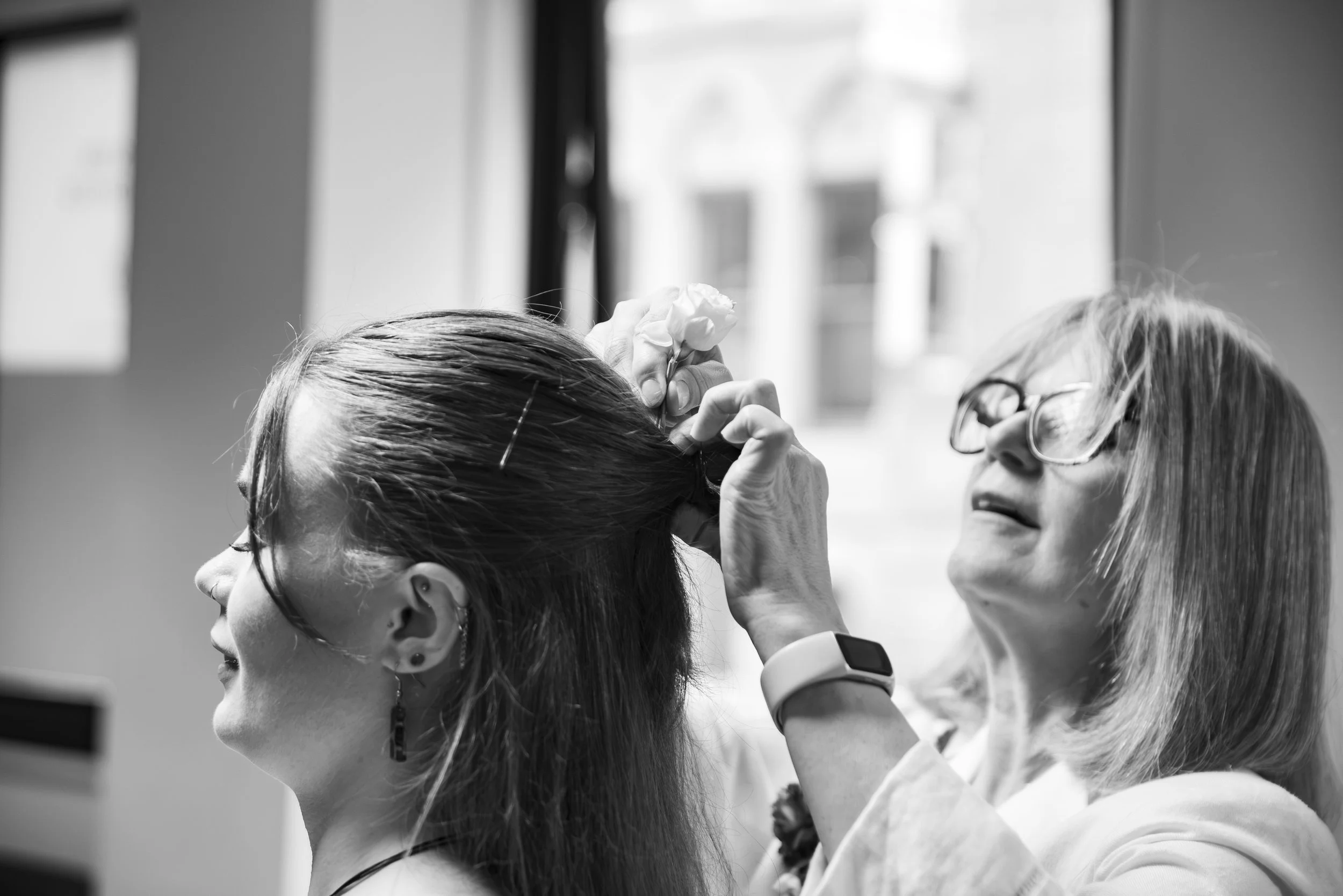 A woman with glasses has her hair styled, while a hairstylist is applying makeup or lipstick to her lips in a salon or hair studio, captured in black and white.