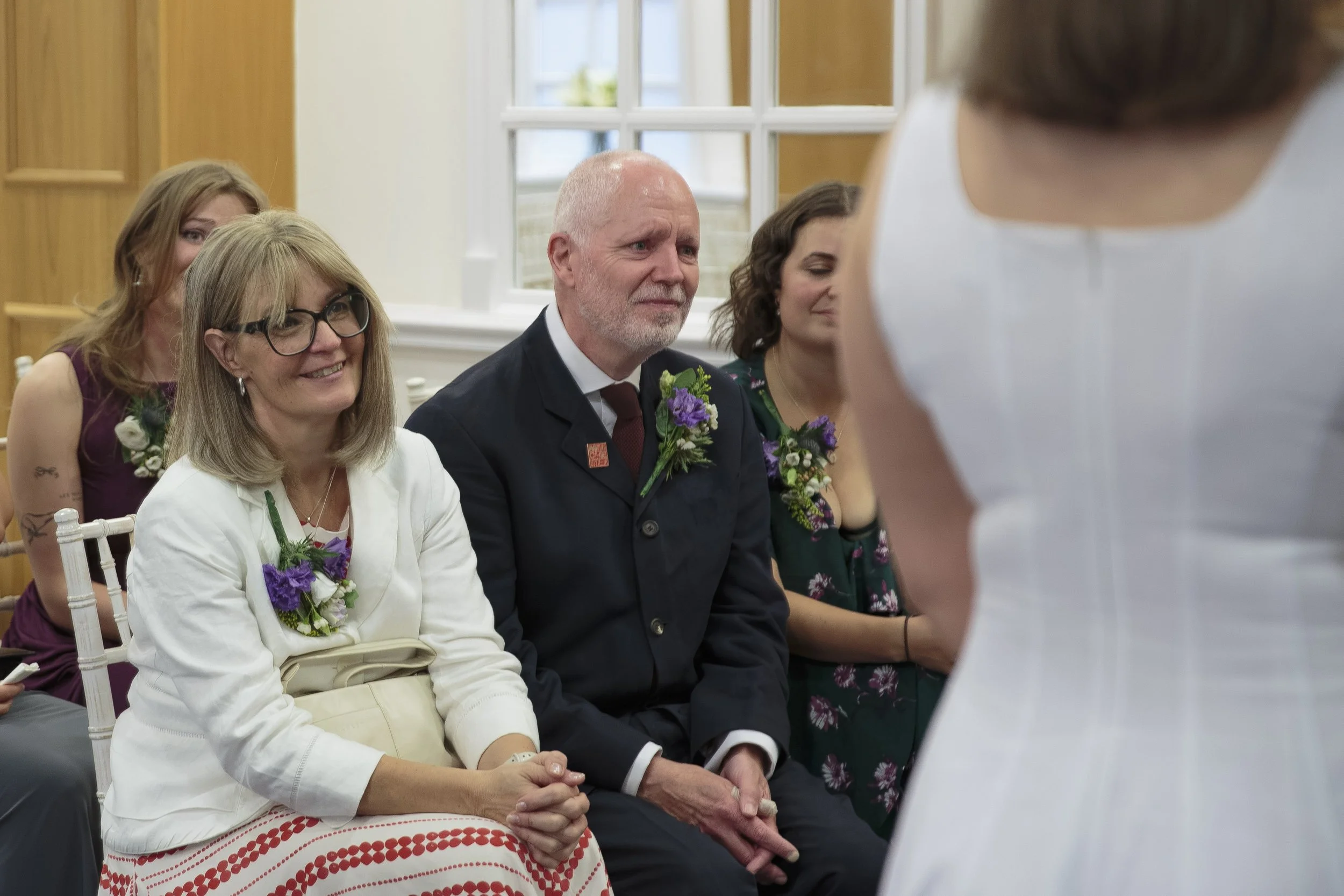 People attending a wedding ceremony, with a woman in white and a man in a dark suit with a boutonniere, sitting and listening to a person in white speaking.
