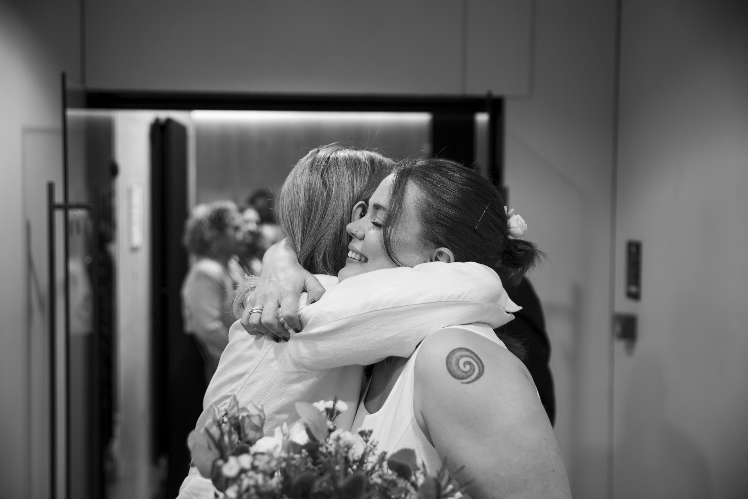 Two women hugging each other in a room, with one holding a bouquet of flowers, seen in black and white.