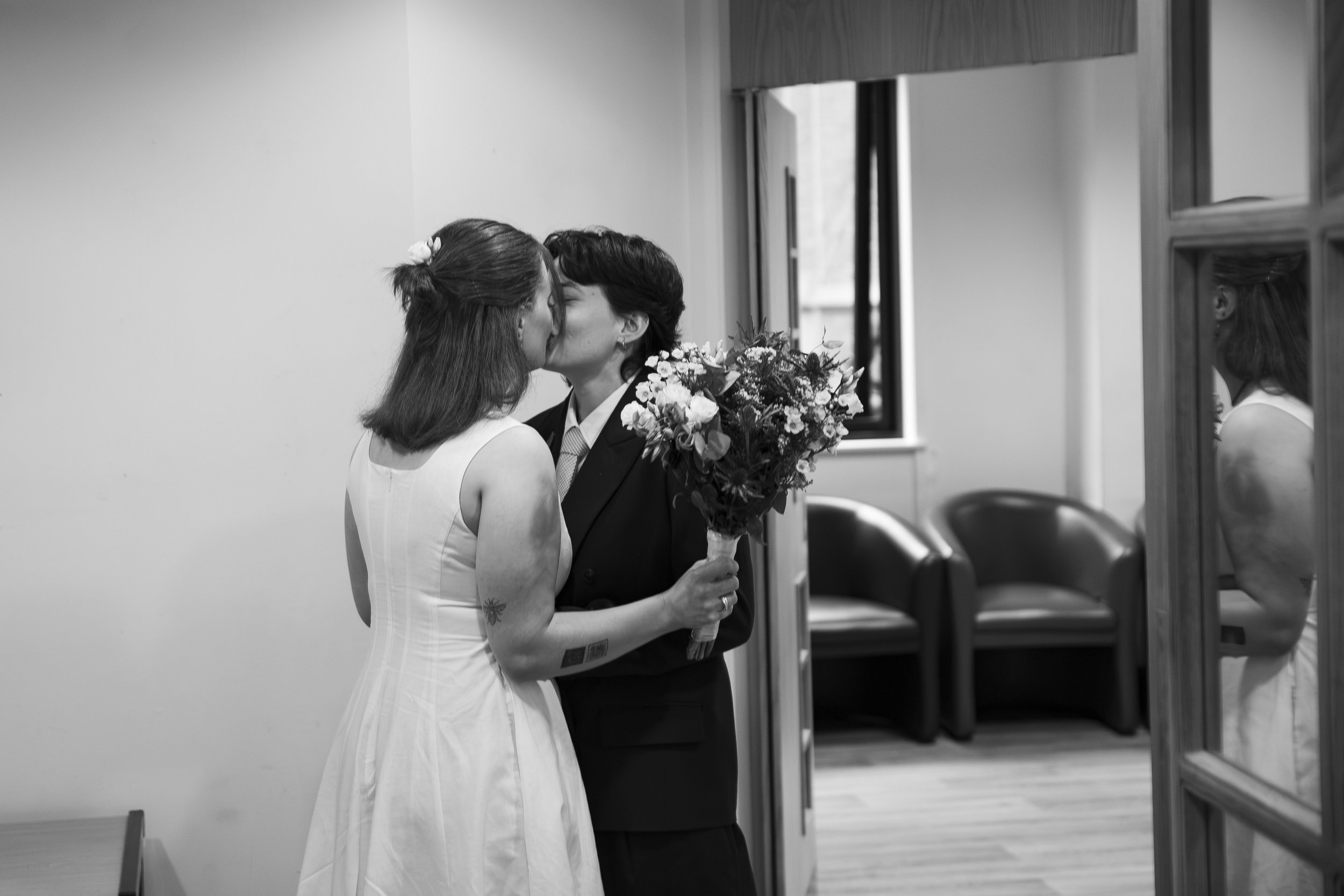 Two women sharing a kiss, one in a wedding dress holding a bouquet, the other in a suit, inside a room with a window and chairs.