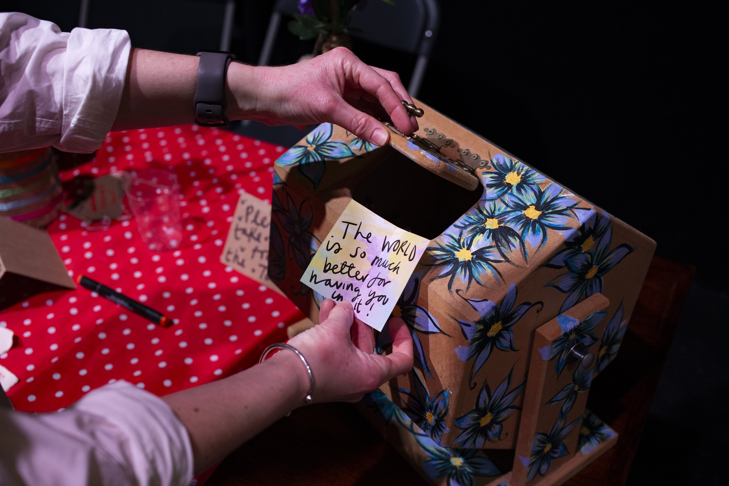 Person wrapping a gift box with floral wrapping paper, while attaching a handwritten note that says, 'The world is so much better for having you in it!' at a table covered with a red polka dot cloth.