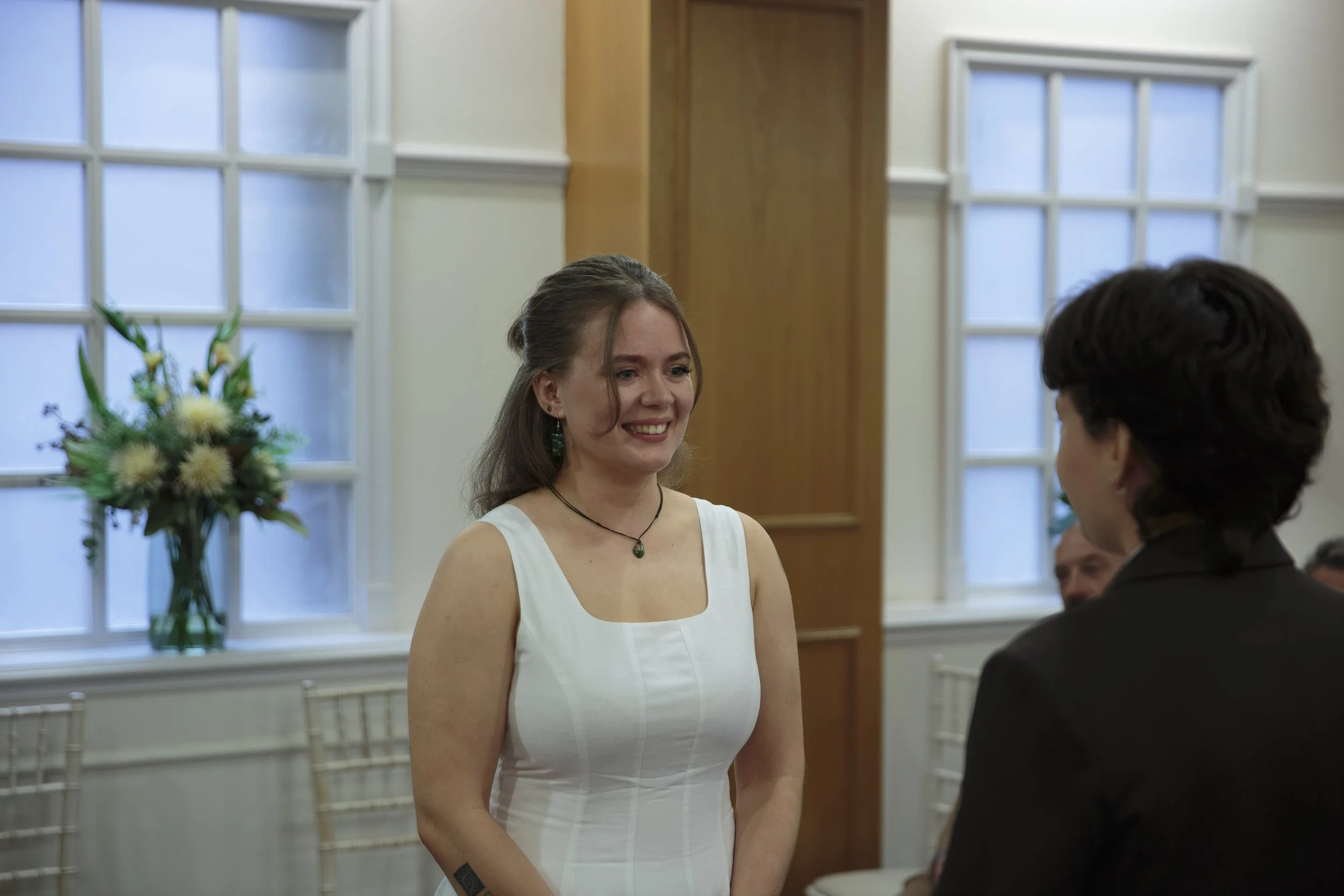 A woman in a white dress and green jewelry smiling while standing during a formal event or ceremony, with people in the background, and a window with a vase of flowers nearby.