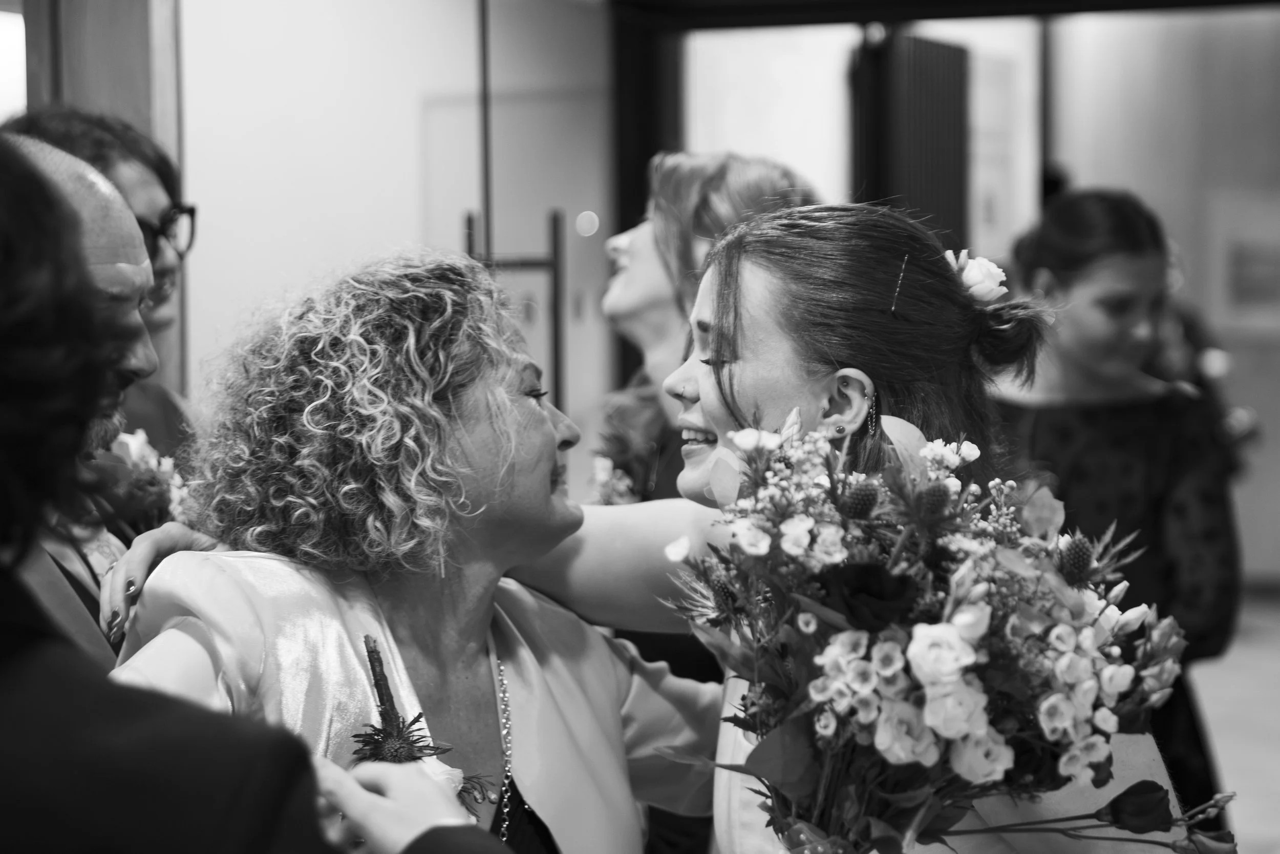 A black and white photo of people at a celebration, possibly a wedding, with women holding bouquets of flowers, smiling and embracing.