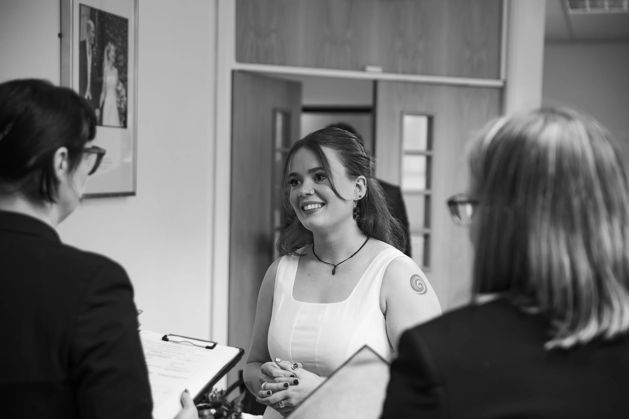 A woman with dark hair, wearing earrings, a necklace, and a sleeveless top, smiling and speaking with three other women in a room, with one holding a clipboard and smiling, engaged in conversation.