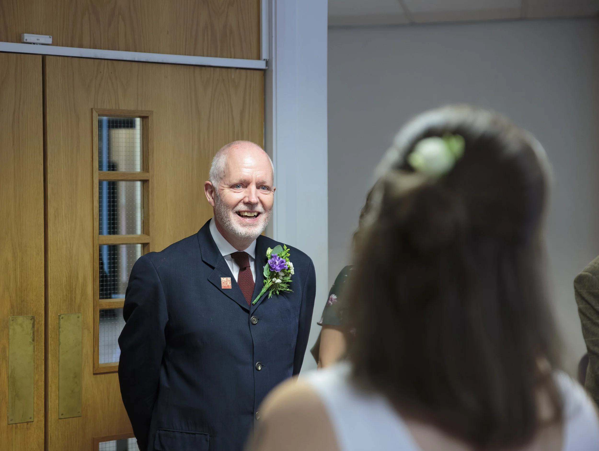 A man with a gray beard and short hair wearing a dark suit and red tie with a floral boutonniere, smiling, standing in front of a wooden door, and talking to a woman.
