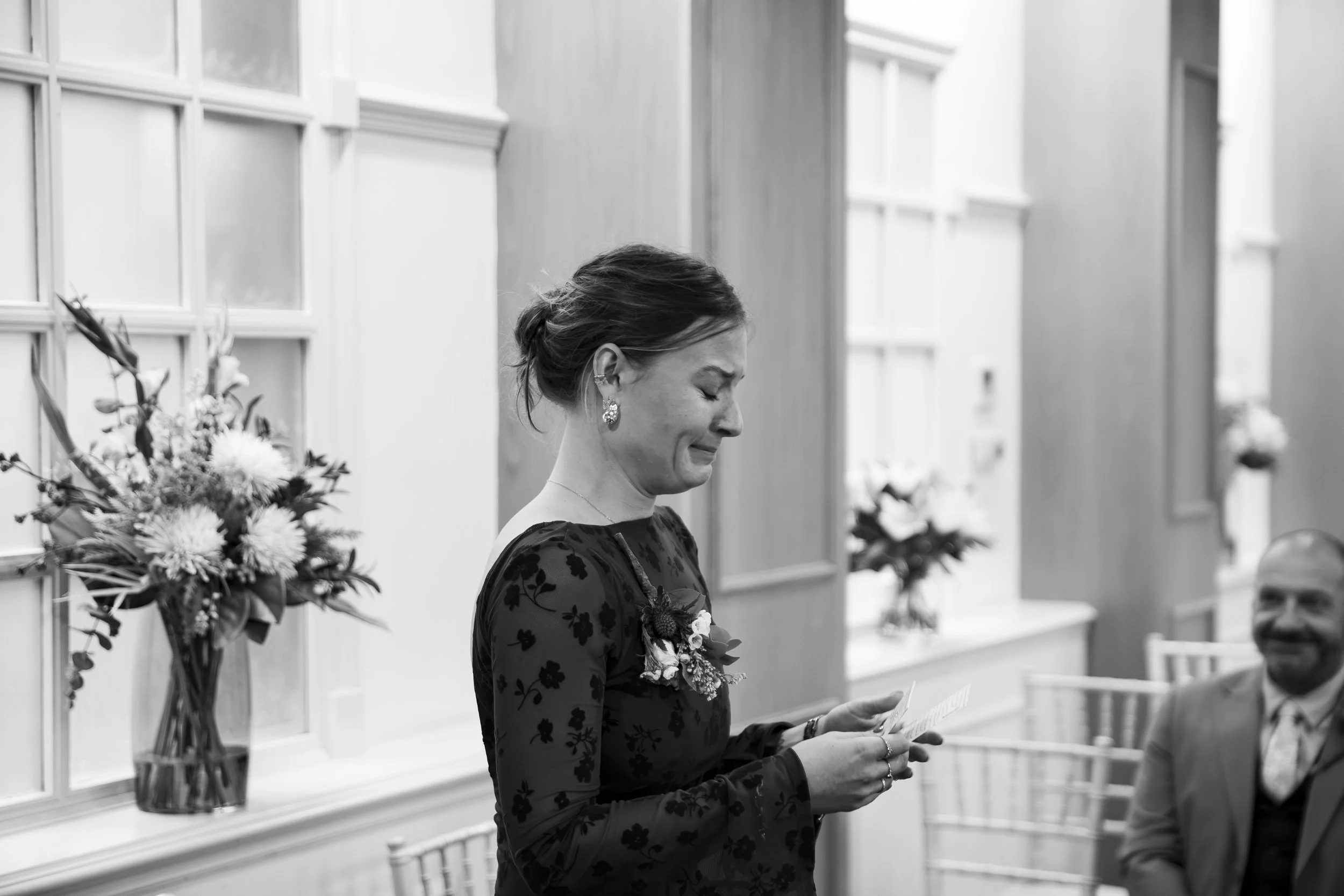 A woman dressed in a black floral dress holding a piece of paper, standing indoors near a window with a floral arrangement visible in the background.