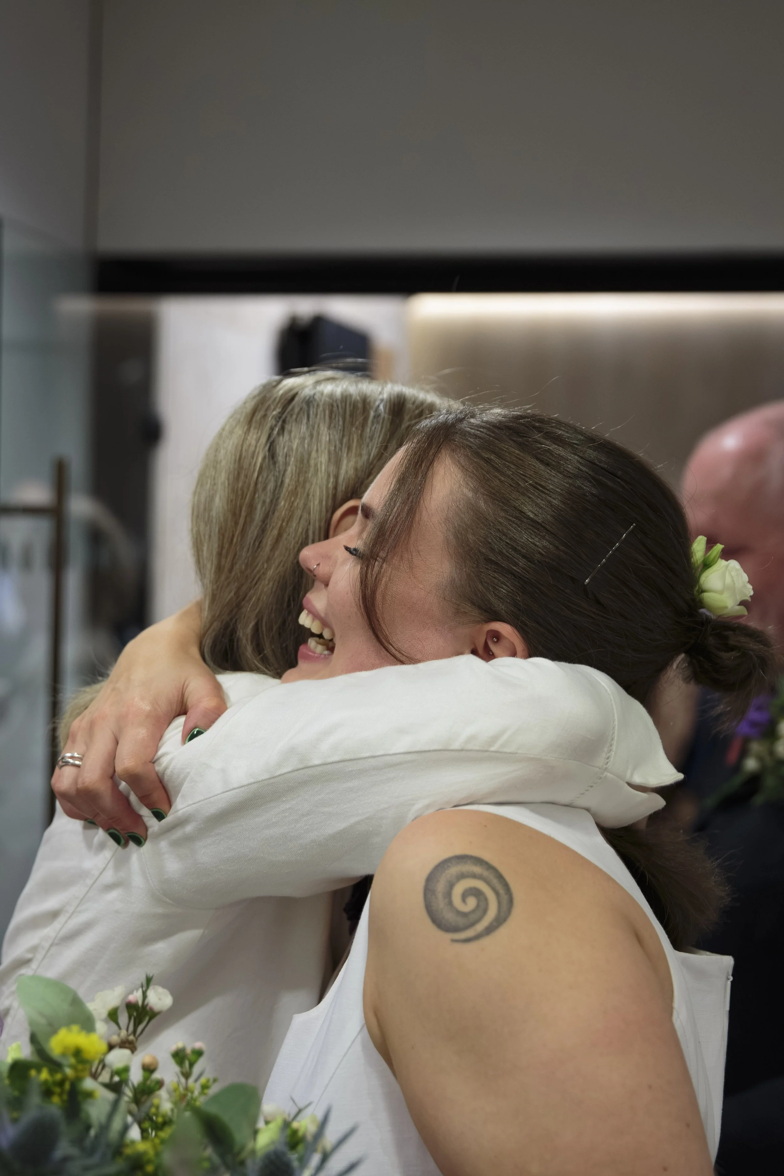 Two women hugging and smiling, one with a spiral tattoo on her arm, in a warm indoor setting.