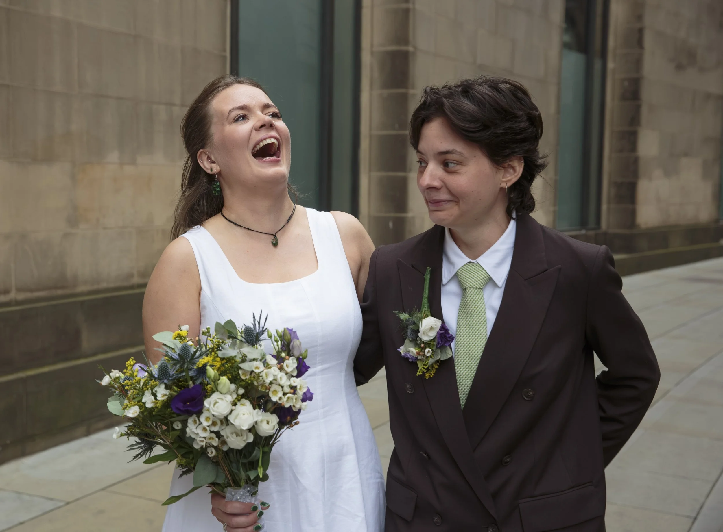Two women standing close together outside, smiling and looking at each other. One is wearing a white dress and holding a large bouquet of colorful flowers, with green earrings and a choker. The other is dressed in a dark suit with a light green tie, 
