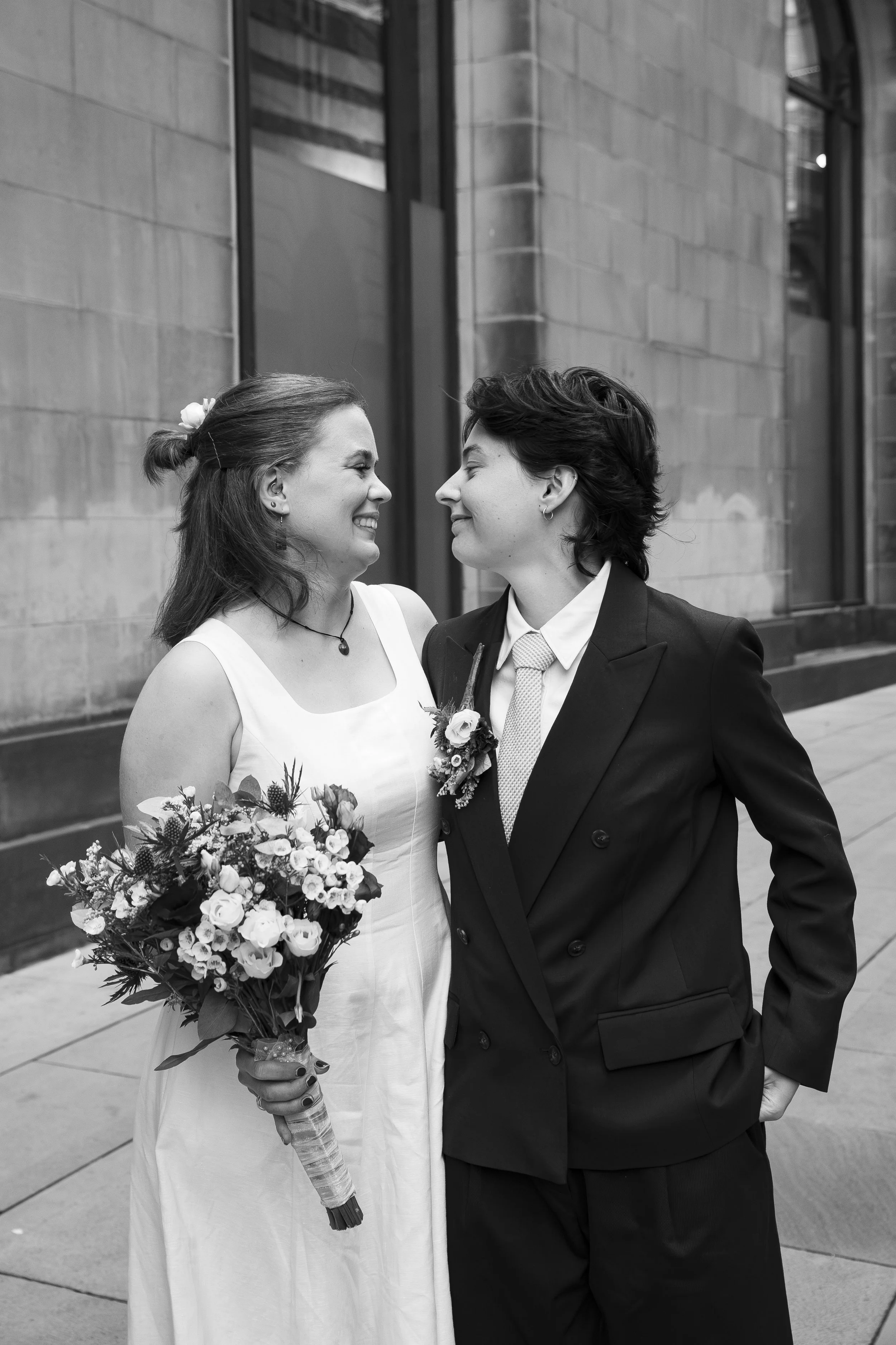 A black and white photograph of two women dressed for a wedding, smiling and leaning close to each other. One woman in a wedding gown is holding a bouquet of flowers, and the other woman in a suit is facing her. They appear to be happy and affectiona
