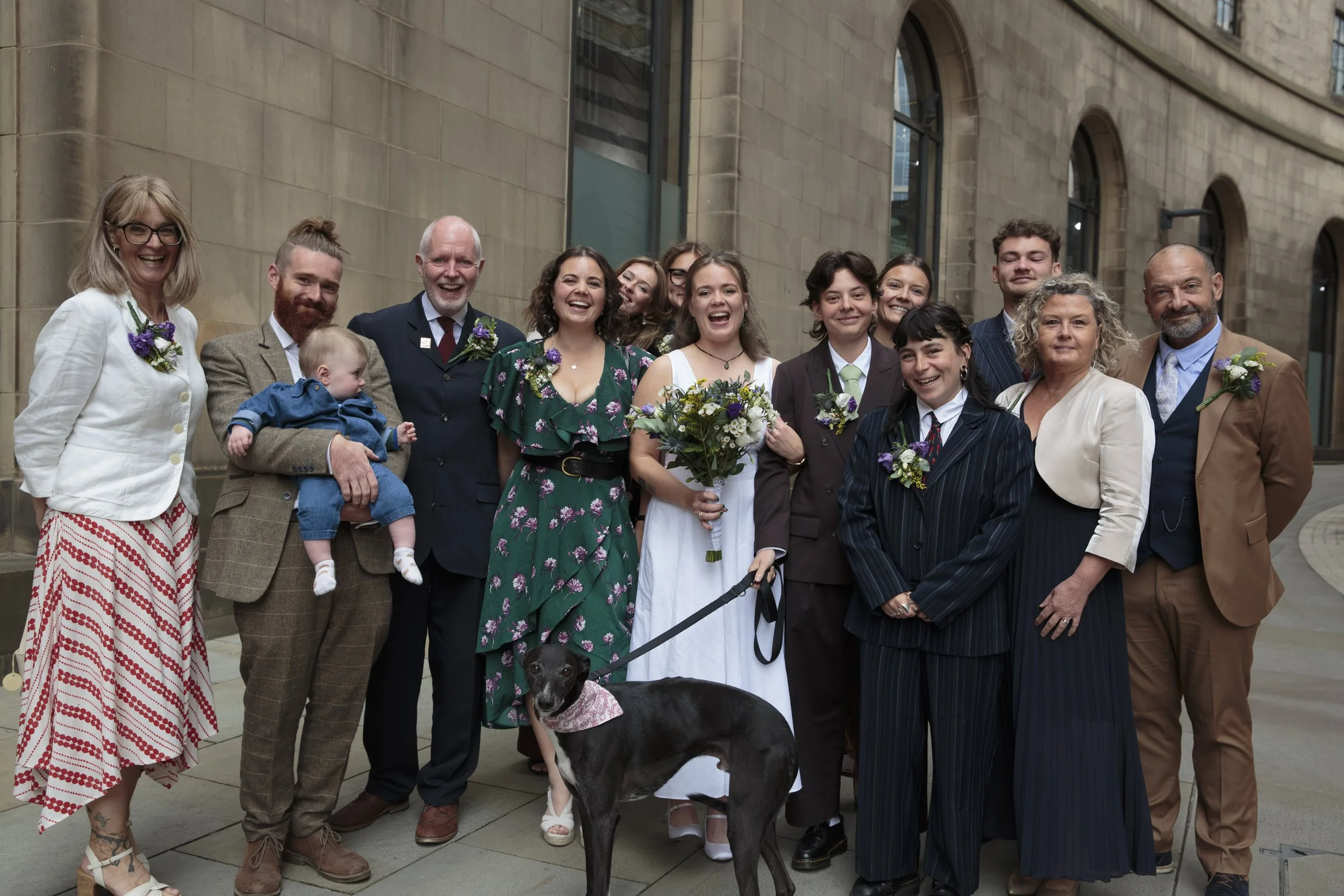 A large group of people standing outside a building, smiling and celebrating. The group includes a woman in a white jacket and patterned skirt, a man holding a small child, various women and men dressed in formal and semi-formal attire, with some wea