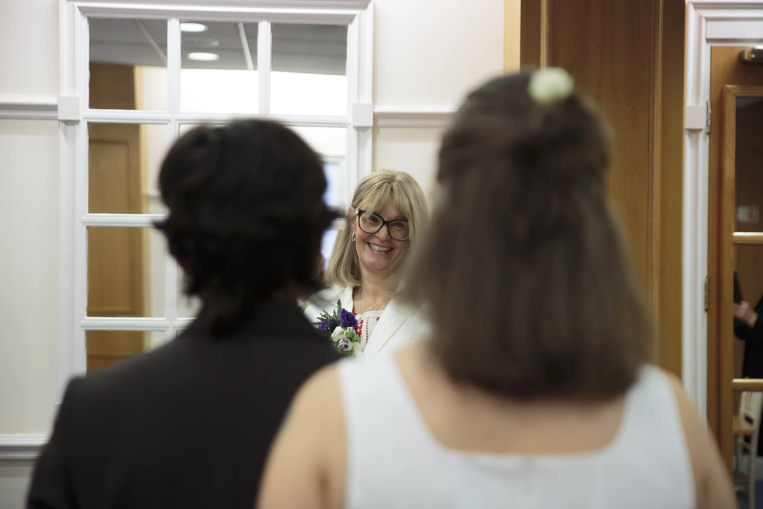 A woman smiling and holding a bouquet, seen through the backs of two other people at what appears to be a gathering or event, with a large mirror and wood-paneled wall in the background.