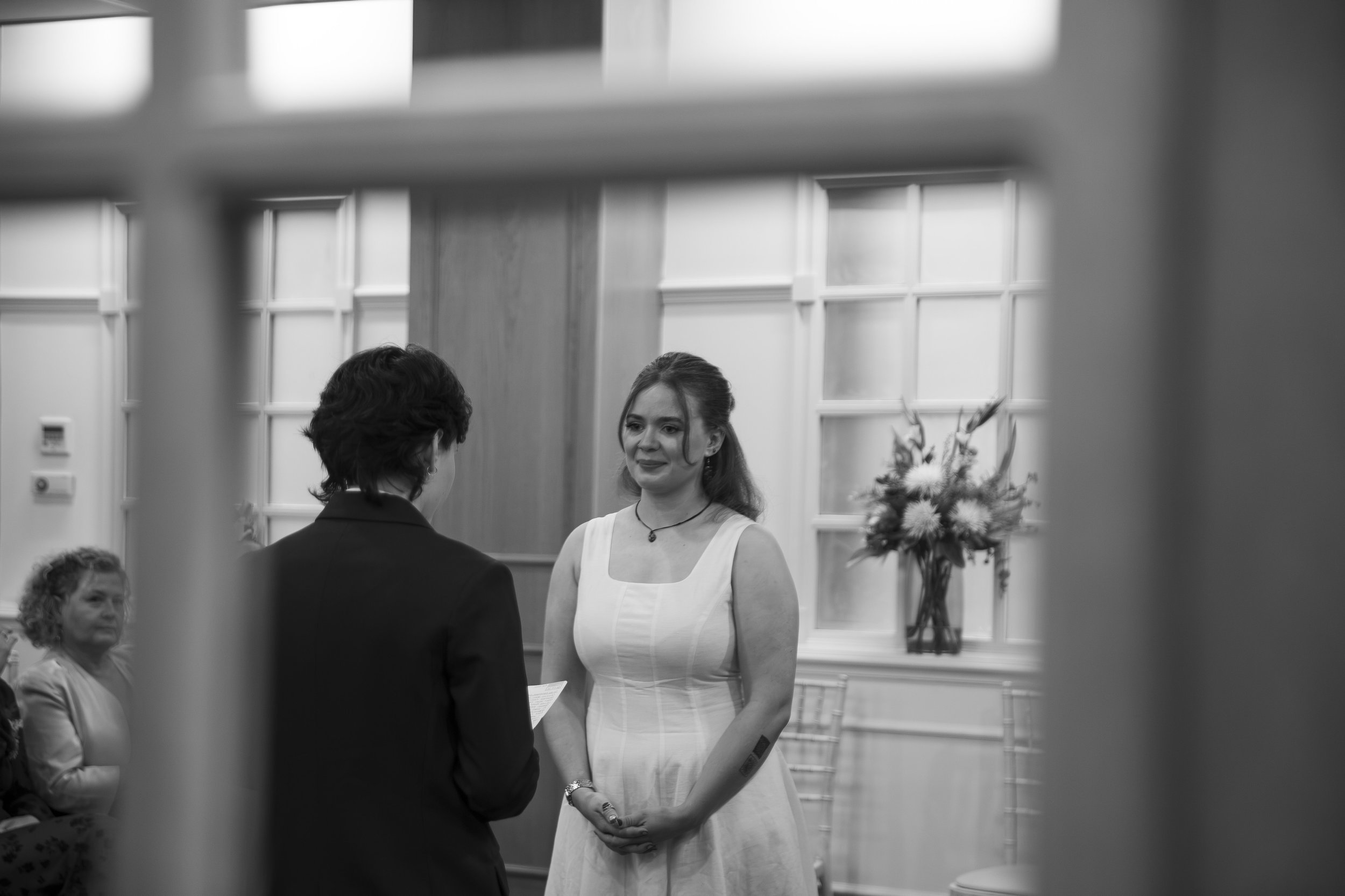 A woman in a white dress and a woman in a black blazer standing face-to-face, with a woman seated in the background to the left, in a room with large windows.