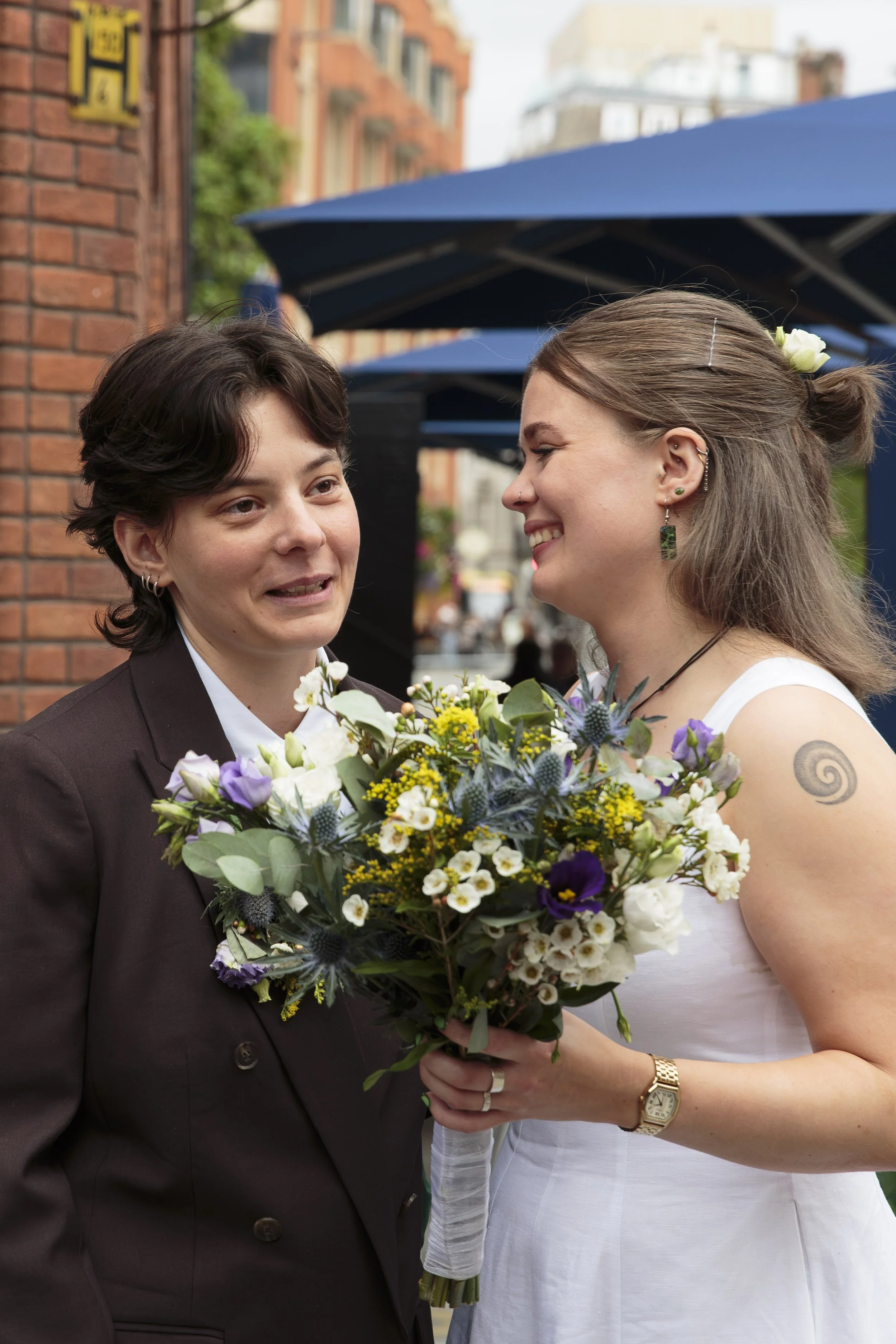 Two women, one dressed in a tuxedo and the other in a wedding dress, are standing outdoors with a bouquet of flowers. They appear to be at a wedding or celebration, smiling and looking at each other. The background includes brick buildings and blue u