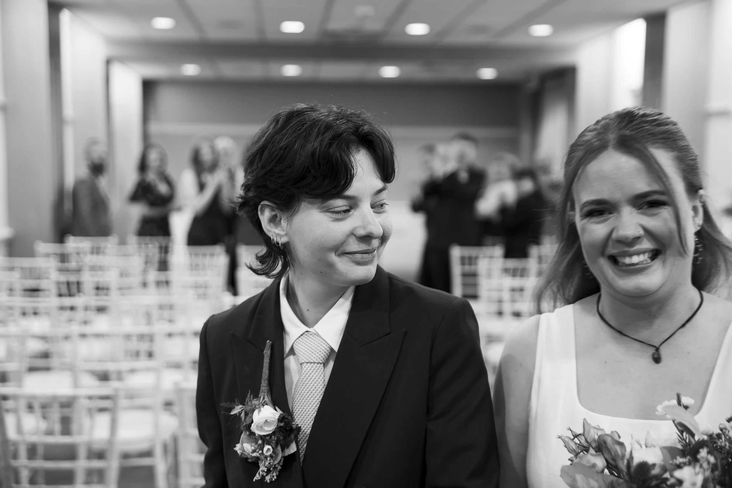 Two women smiling, dressed in formal attire, at a wedding or celebration, with blurred guests and chairs in the background.
