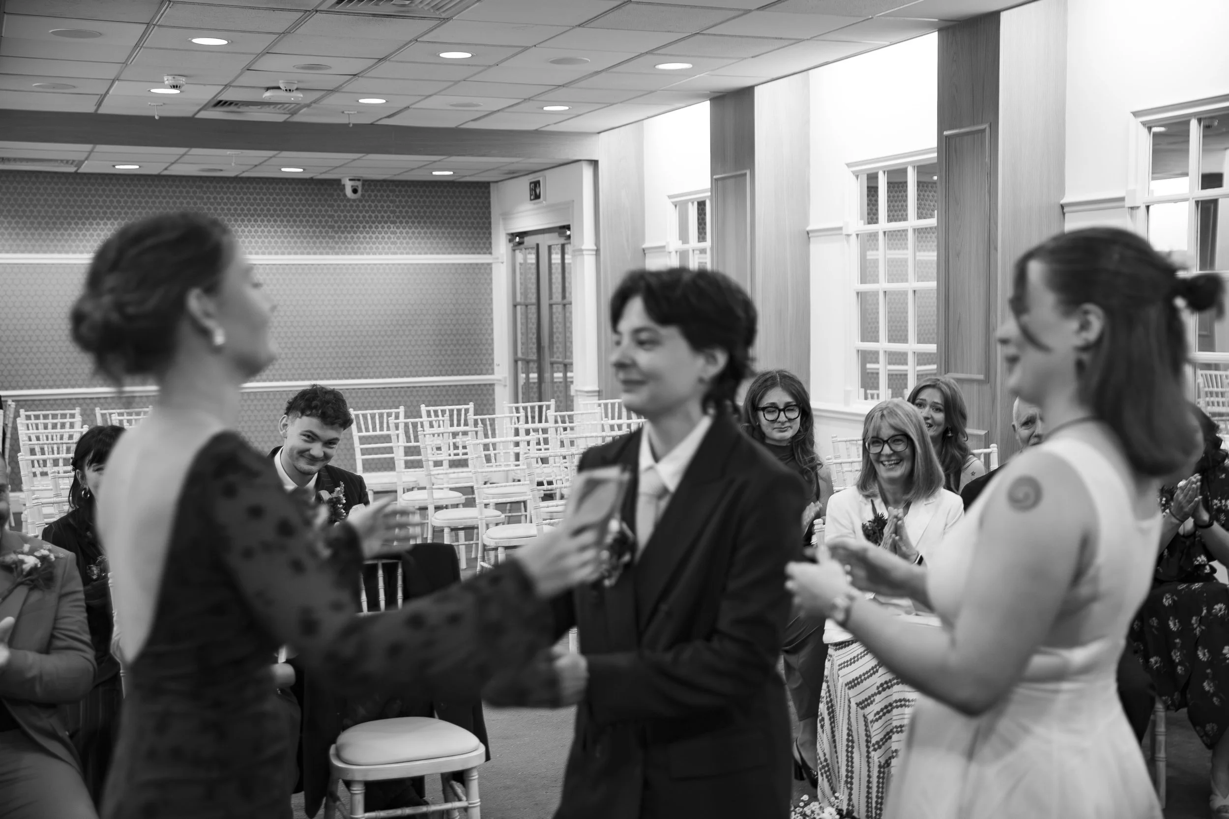 People attending a wedding ceremony in an indoor venue, with two women in the foreground holding hands and exchanging vows, while others sit and watch in the background.