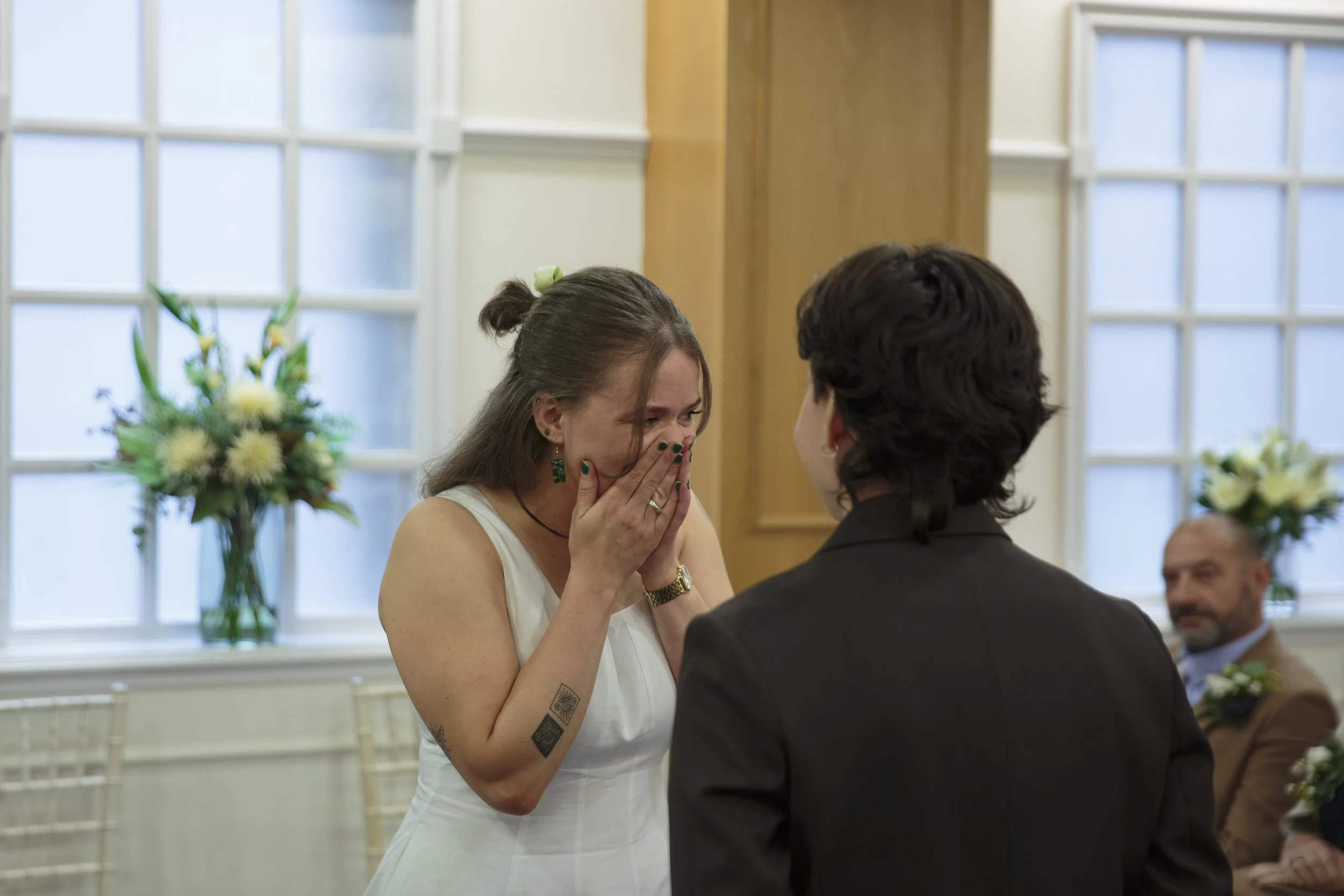 A woman with long dark hair, wearing a white dress, is crying and covering her face with her hands during what appears to be a wedding ceremony. She is facing a man dressed in a dark suit. There are flowers in the background along with large windows 