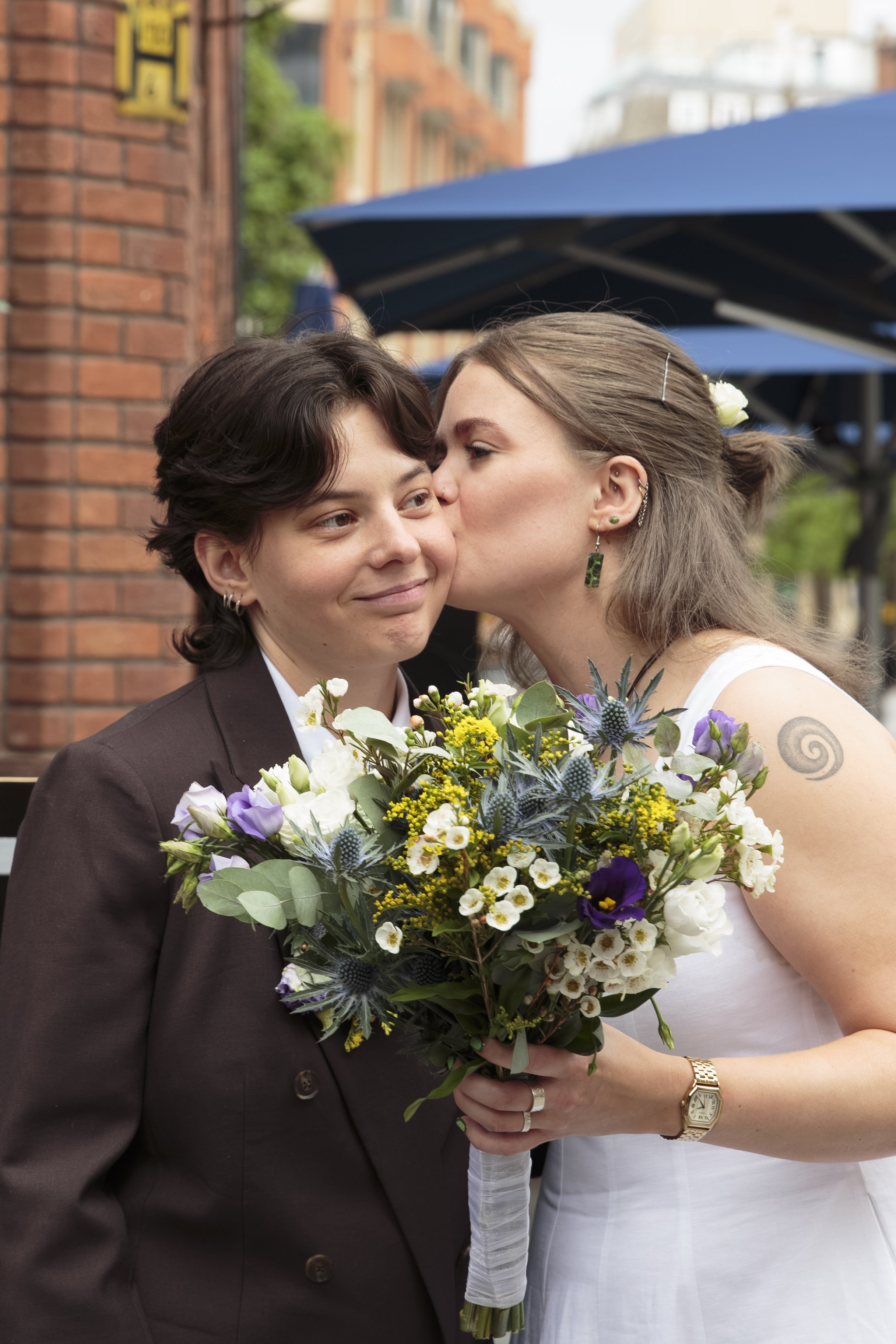 A same-sex couple by a red brick building, with one person kissing the other's cheek while holding a bouquet of mixed flowers. The person receiving the kiss is wearing a white dress and various earrings, and has a tattoo on their arm. The person givi