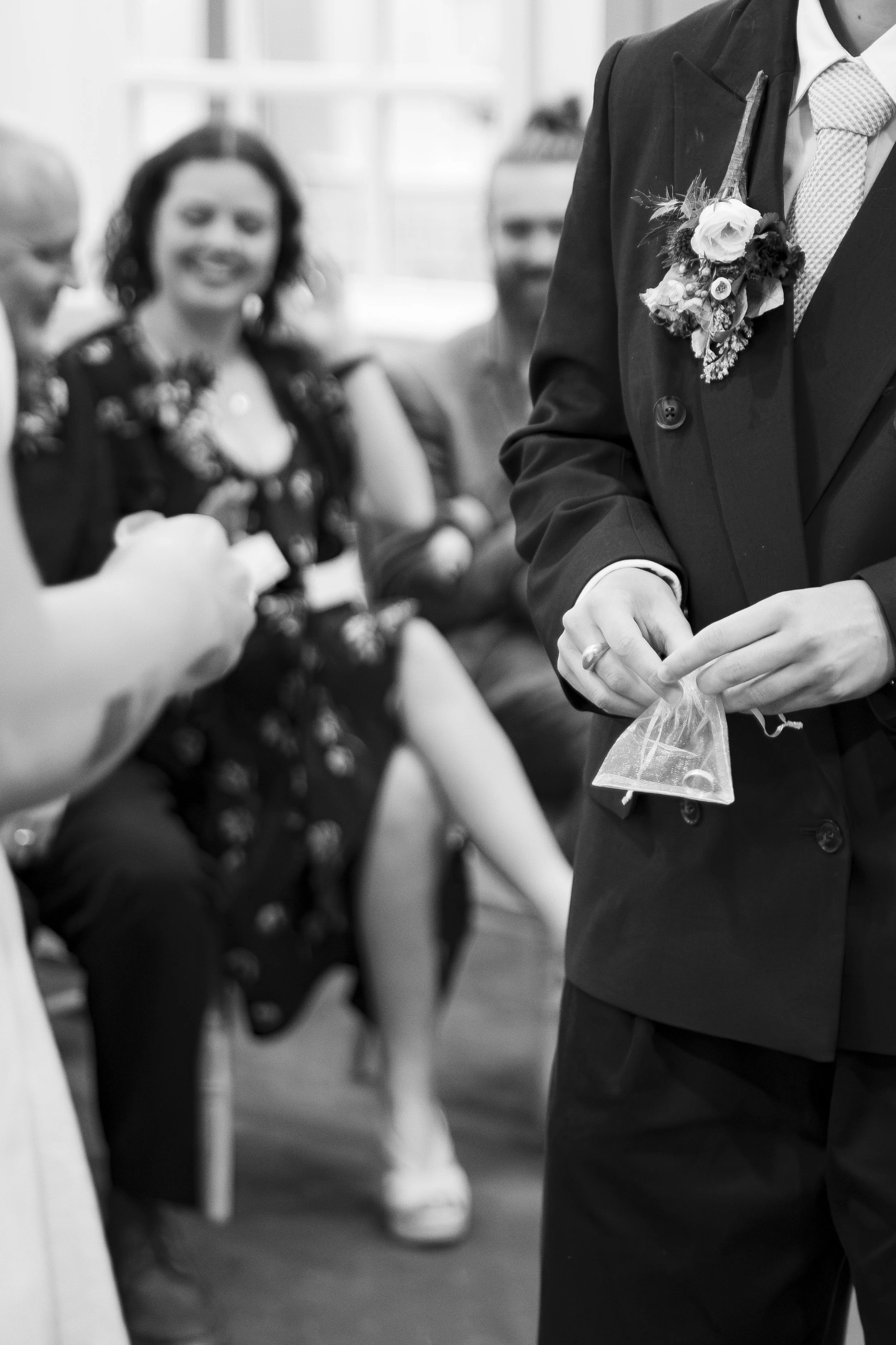 A man in a suit and tie with a boutonniere on his jacket holding a small clear plastic bag, with two women in the background looking on during what appears to be a social event or gathering.