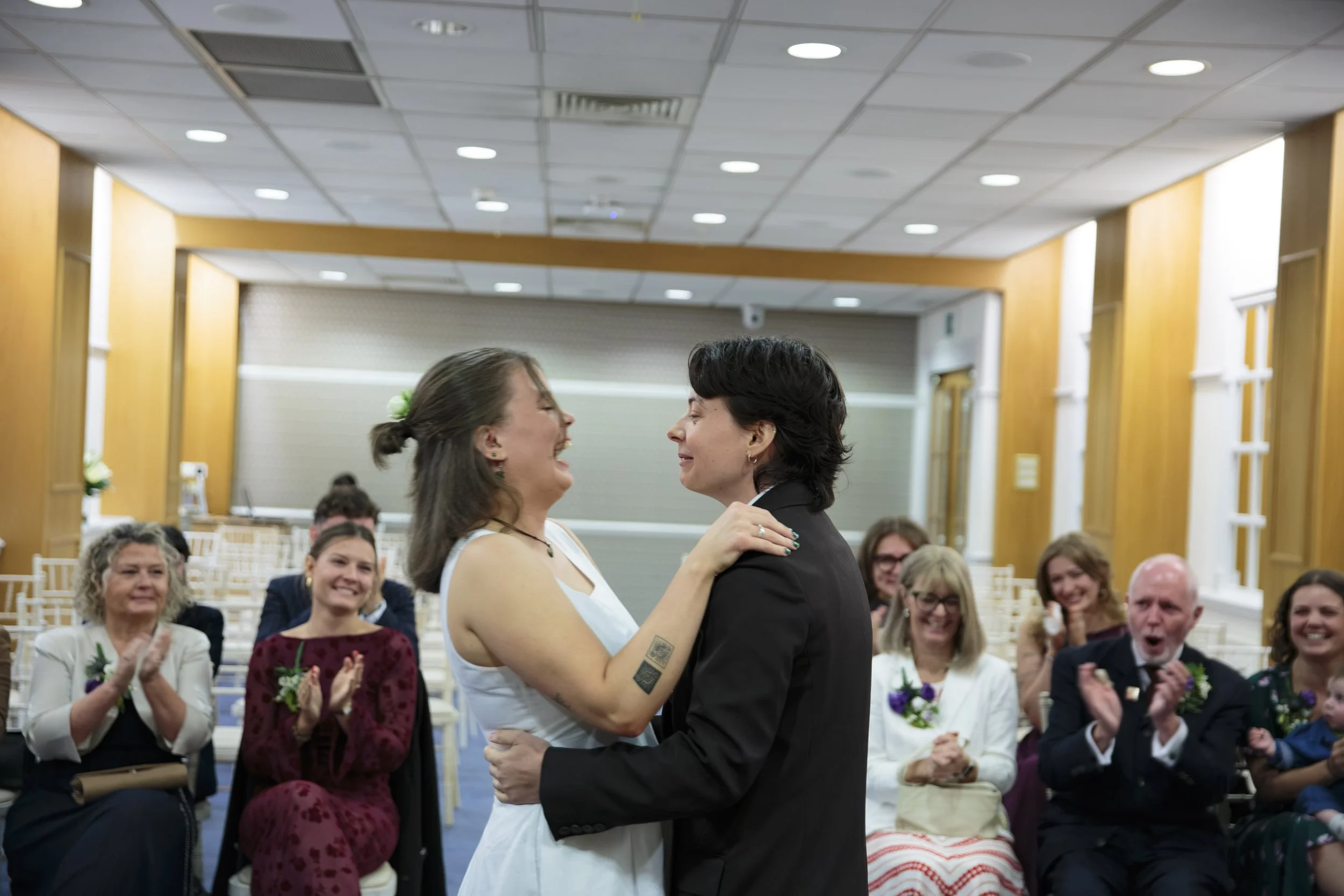 Two women dancing together at a wedding reception, surrounded by seated guests who are clapping and smiling.