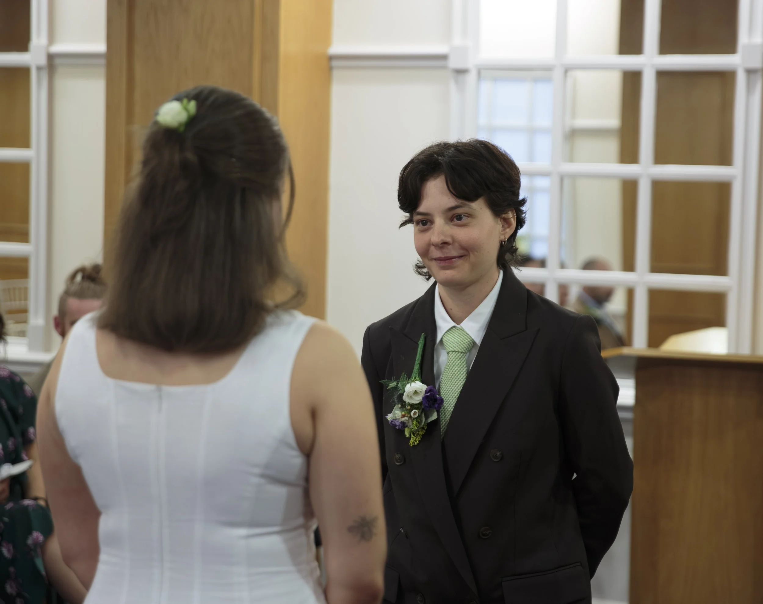 Young couple at a wedding, the woman in a white dress with a flower in her hair, and the man in a suit with a boutonniere, facing each other in a bright, decorated venue.