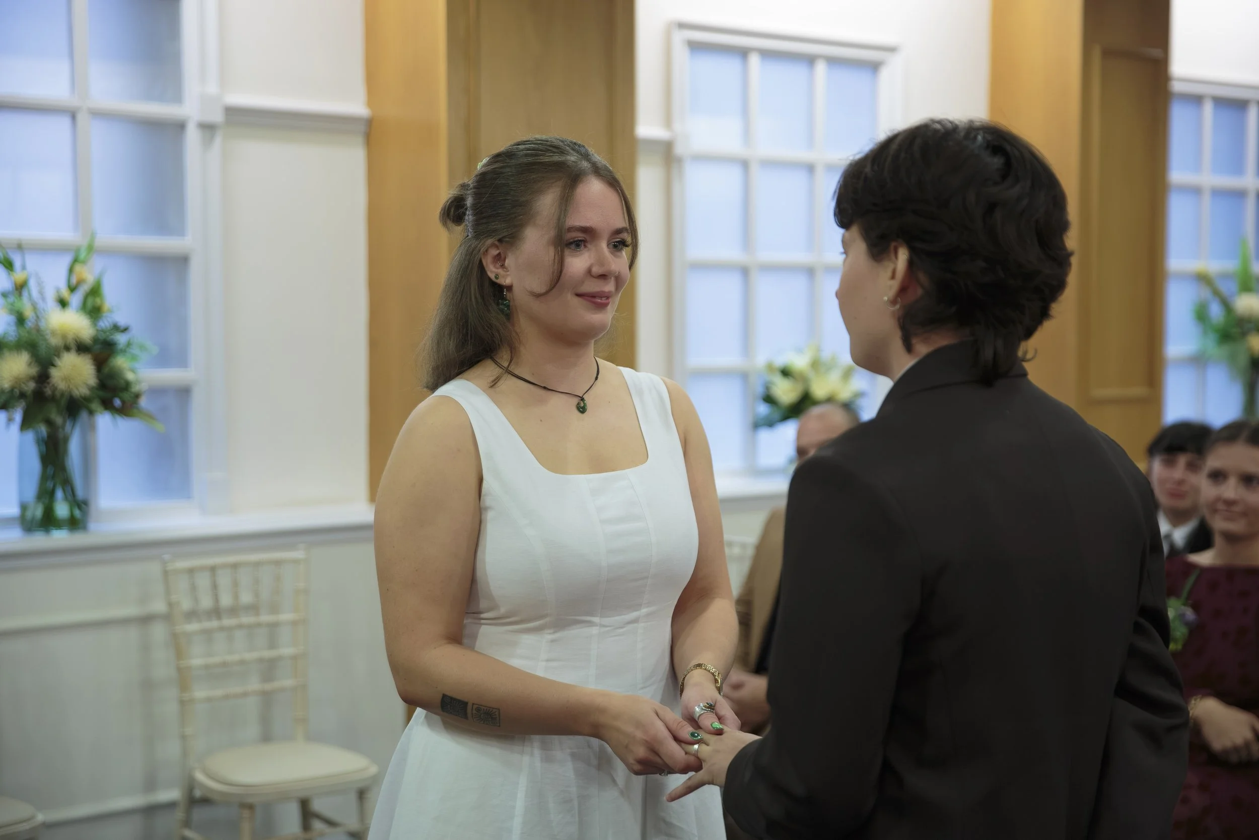 A woman in a white dress and a man in a black suit are holding hands and looking at each other during a wedding ceremony. There are people seated in the background, a bouquet of flowers on a table, and large windows with natural light.