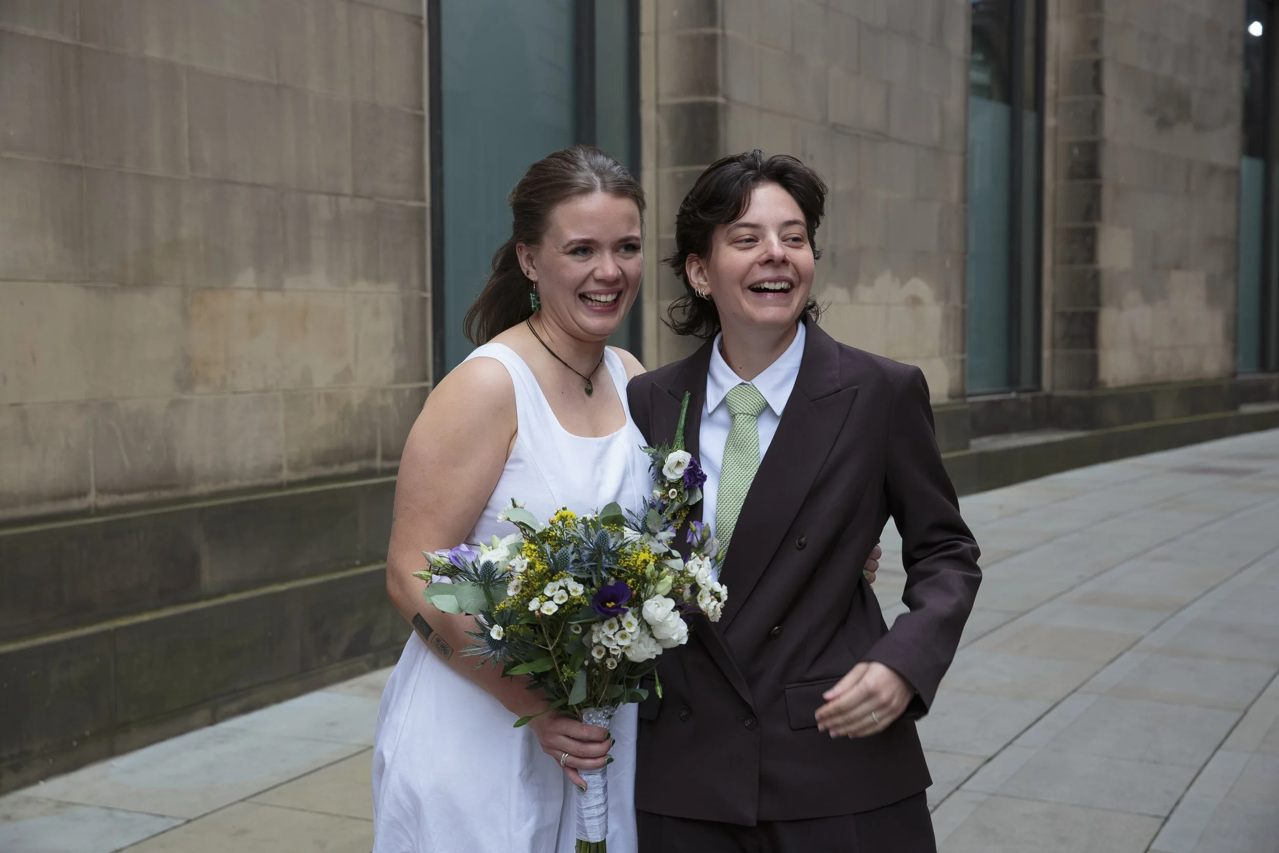 Two women smiling and walking together outdoors, one wearing a white dress holding a bouquet of flowers, the other in a dark suit and green tie.