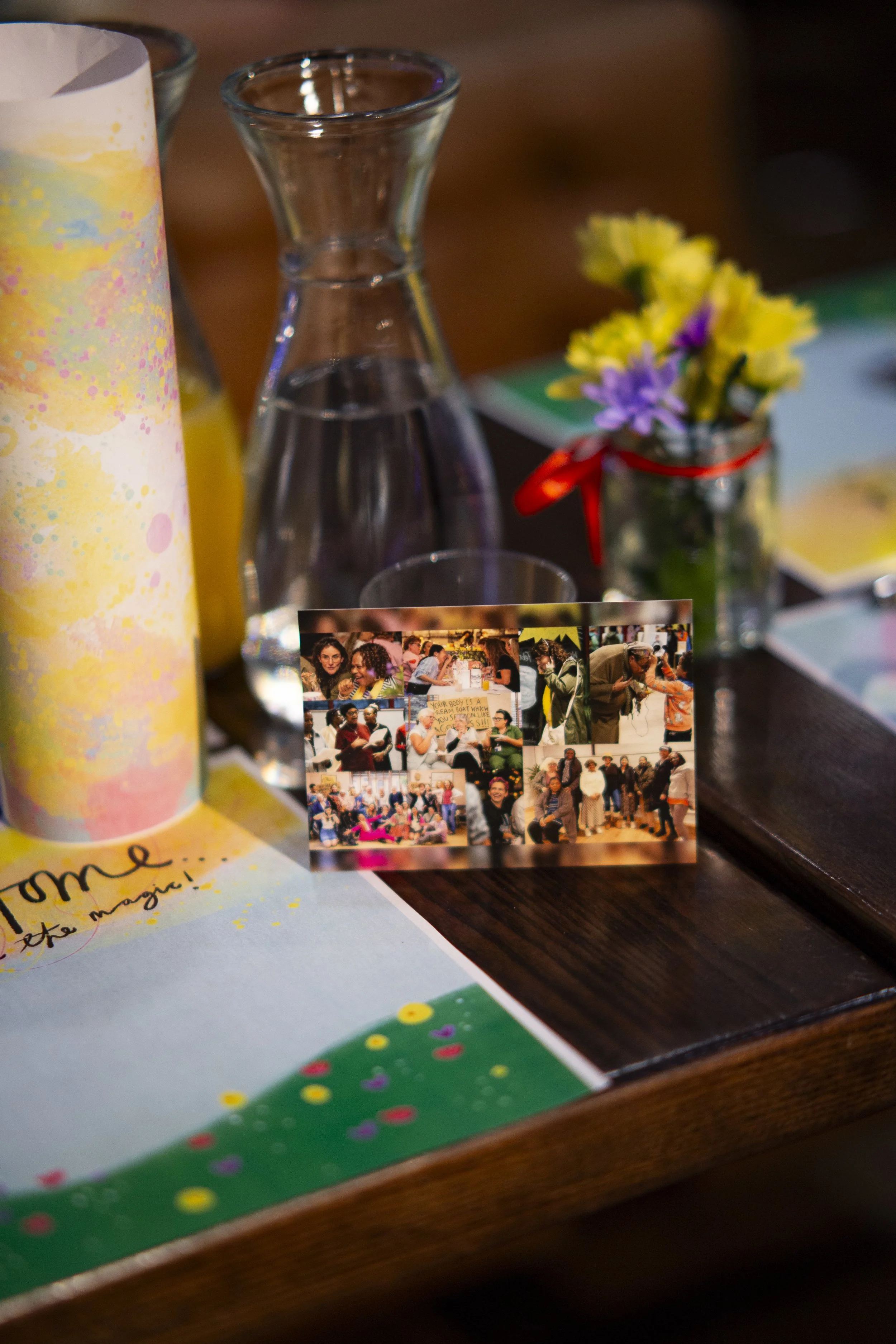 Close-up of a table with a small bouquet of yellow and purple flowers in a jar, a glass water pitcher, a small photo collage, and colorful paper with handwritten text.