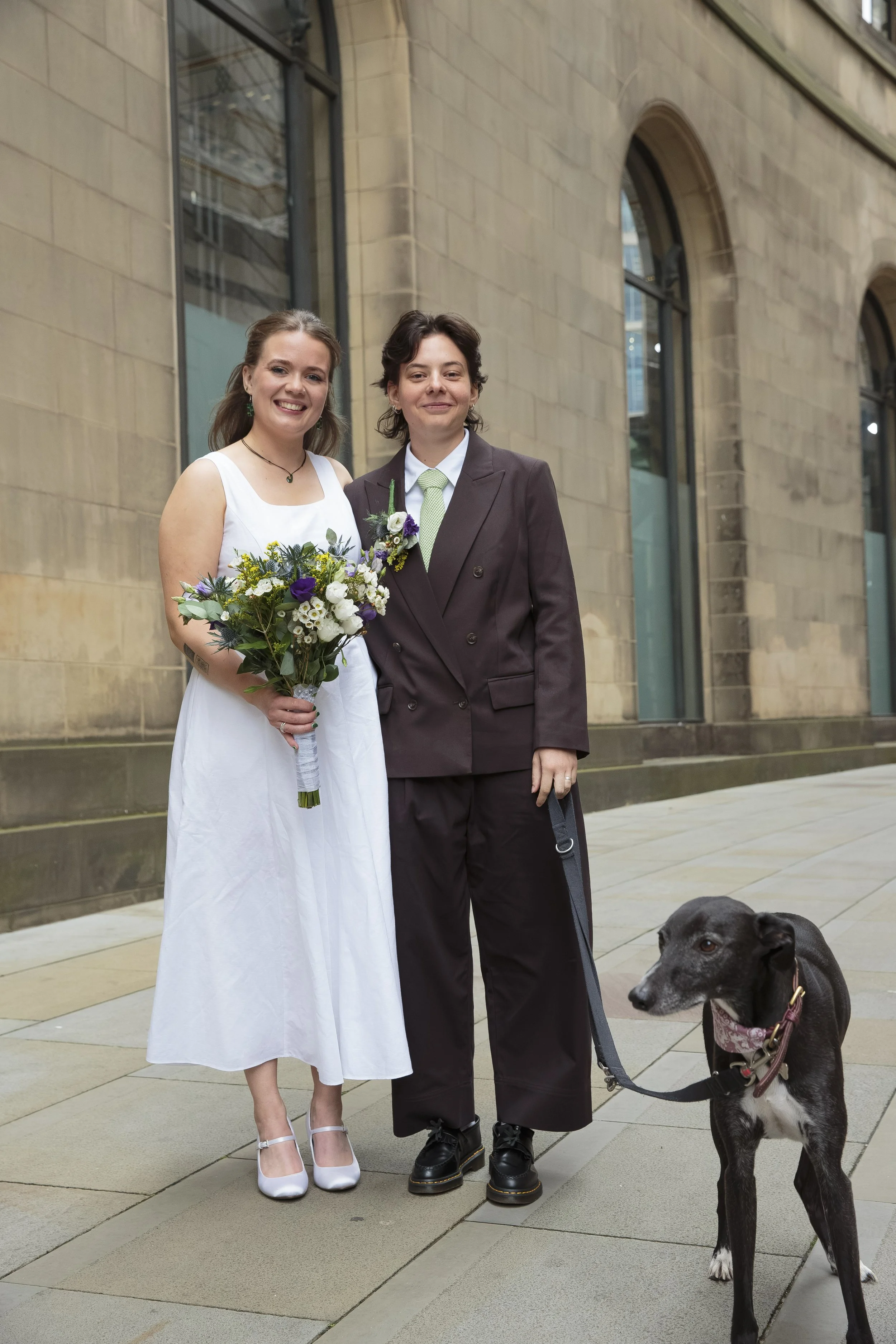 Two women standing outside a stone building, one in a white dress holding a bouquet of flowers, the other in a dark suit with a green tie, and a black dog on a leash nearby.