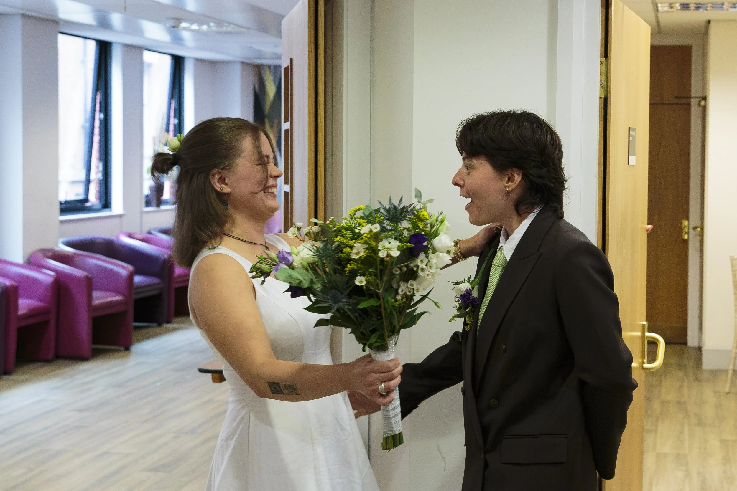 A woman in a white dress and a woman in a black suit holding a bouquet of flowers and hugging in a room with pink chairs and windows.