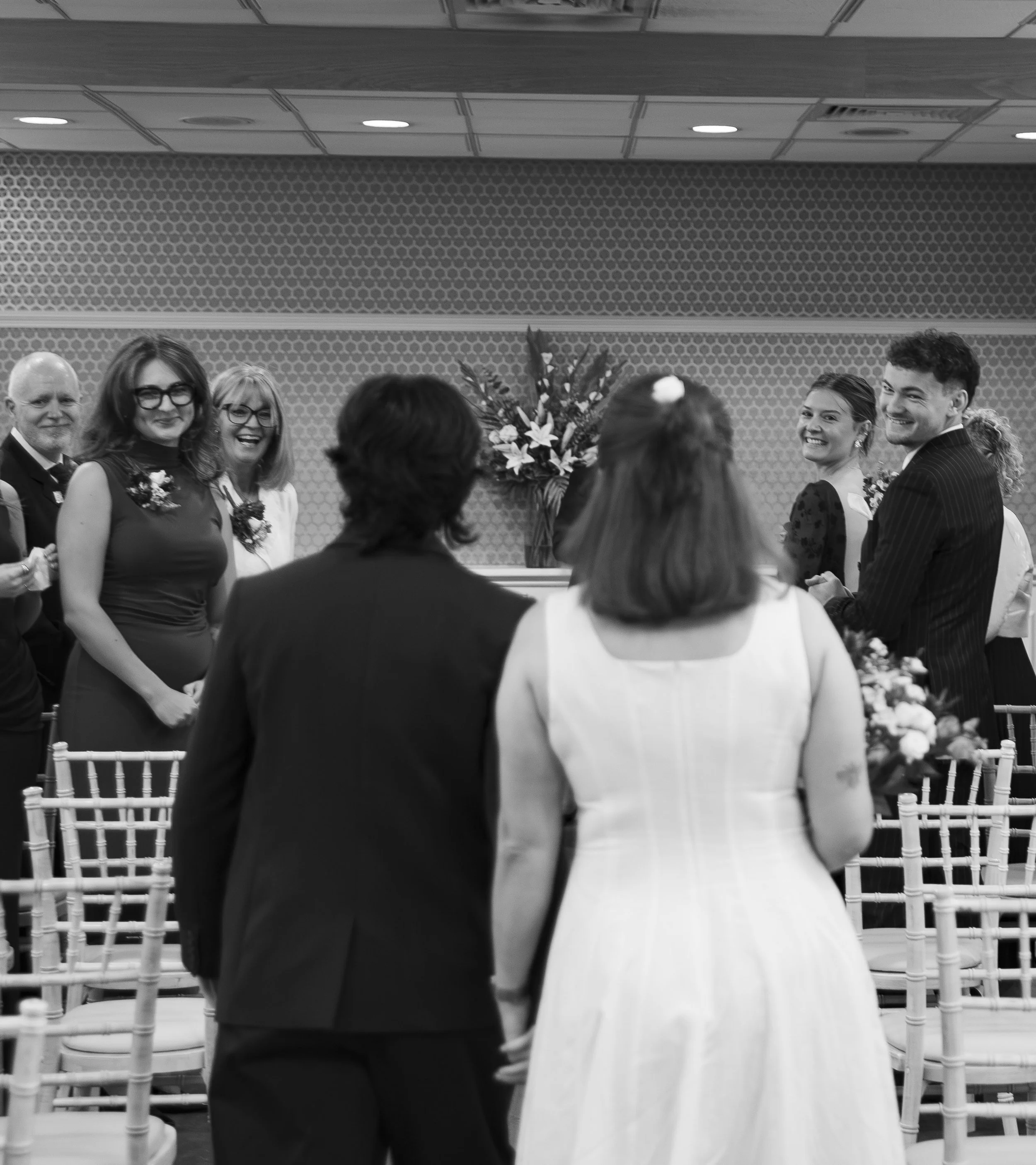 People gathered at a formal event, standing in a circle, with flowers and chairs visible in the setting, likely a wedding or reception.