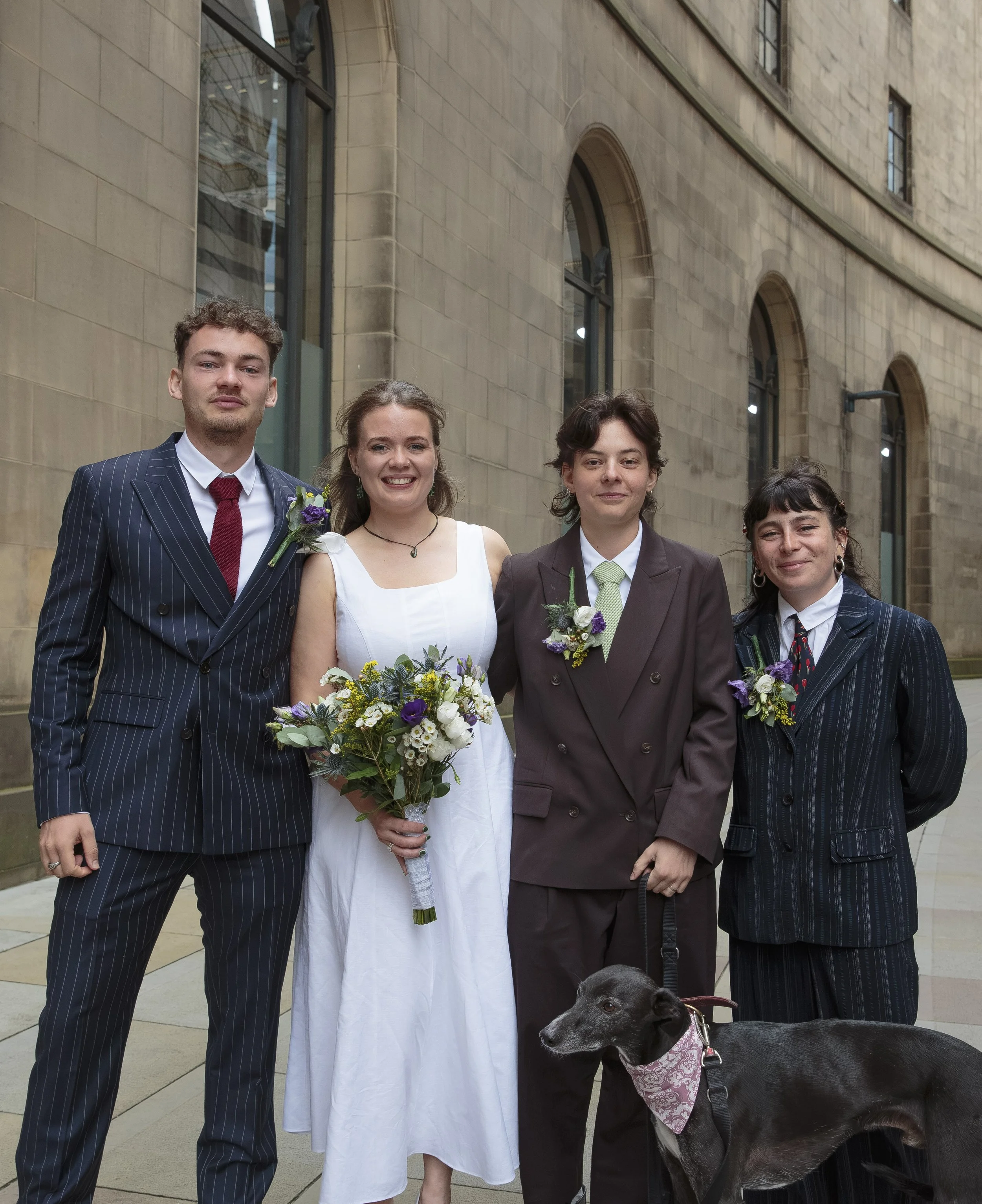 Four people, three women and one man, are standing outdoors in front of a stone building. The woman in the center is wearing a white dress and holding a bouquet of flowers. The others are dressed in suits, and a black dog with a pink bandana is in fr