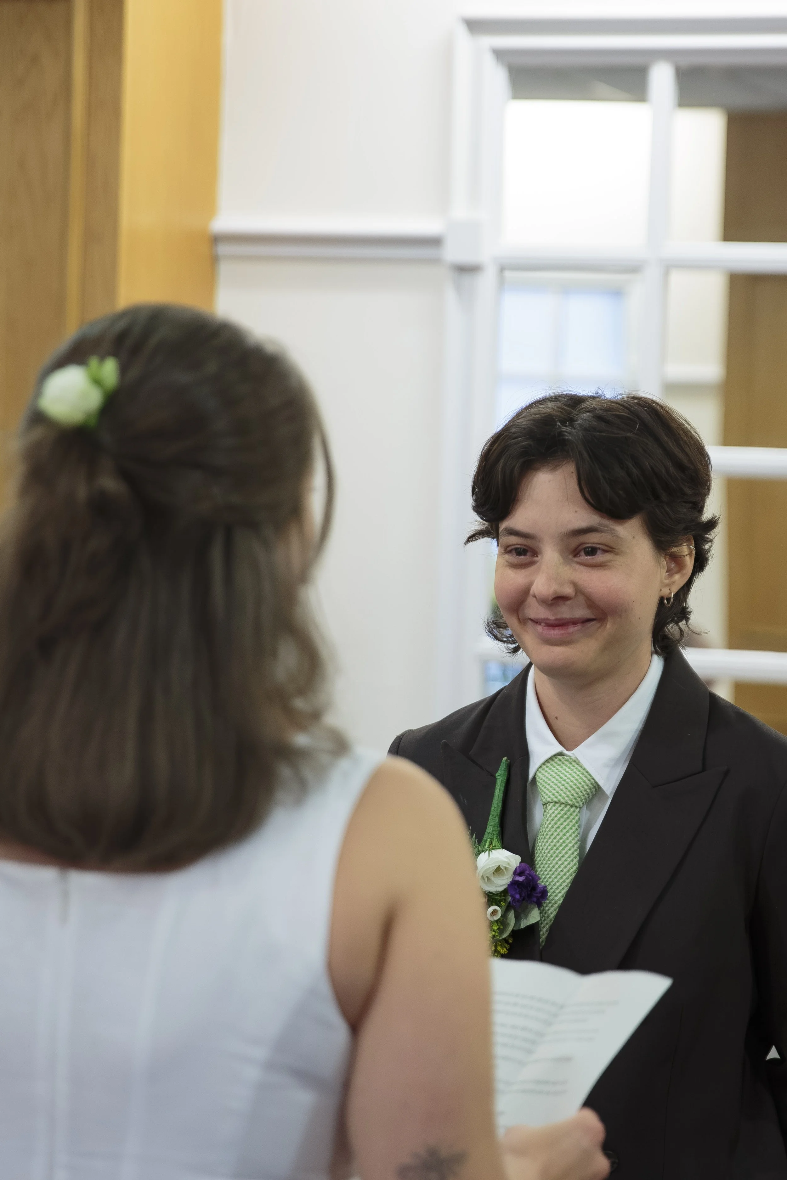 A woman wearing a white dress with a green hair accessory is standing in front of a man dressed in a black suit with a green tie, during a wedding ceremony. The man is smiling and holding a paper, with a white flower corsage on his lapel.
