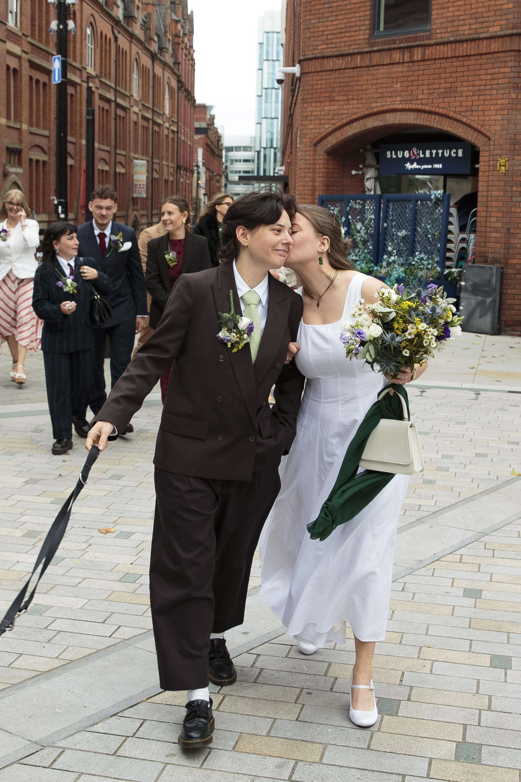 A couple dressed in wedding attire share a kiss while walking on a city street, surrounded by friends and family.
