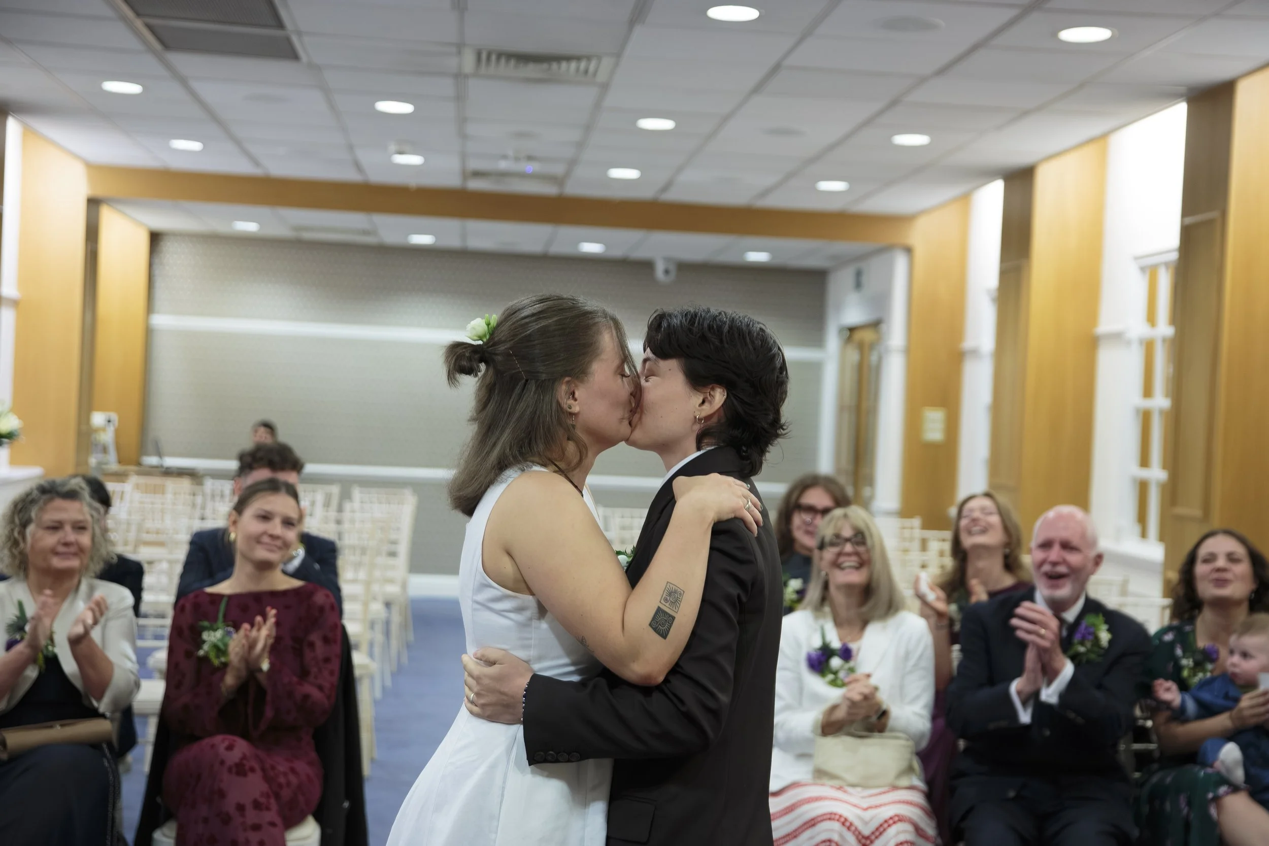 Two women, one in a white dress and the other in a black tuxedo, are kissing at a wedding ceremony. They are surrounded by smiling guests seated and clapping in a banquet hall with wooden paneling and a high ceiling.