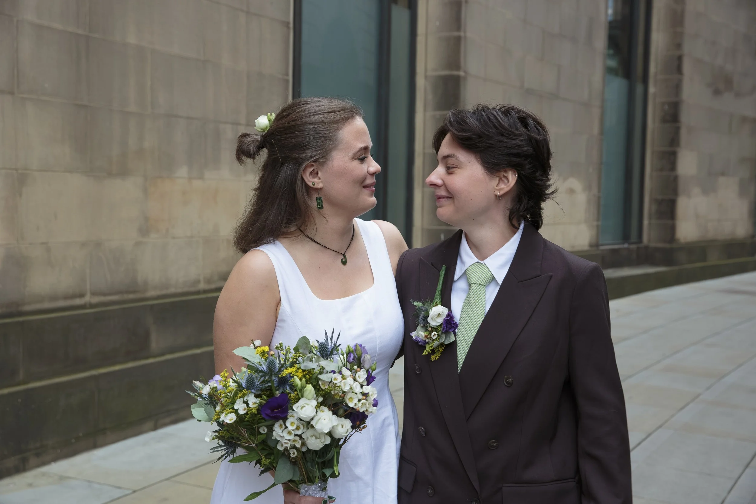 A bride and groom are close together, smiling and touching foreheads outside a stone building. The bride has brown hair tied with a flower and wears a white dress, holding a colorful bouquet. The groom has dark hair, wears a dark suit with a green ti
