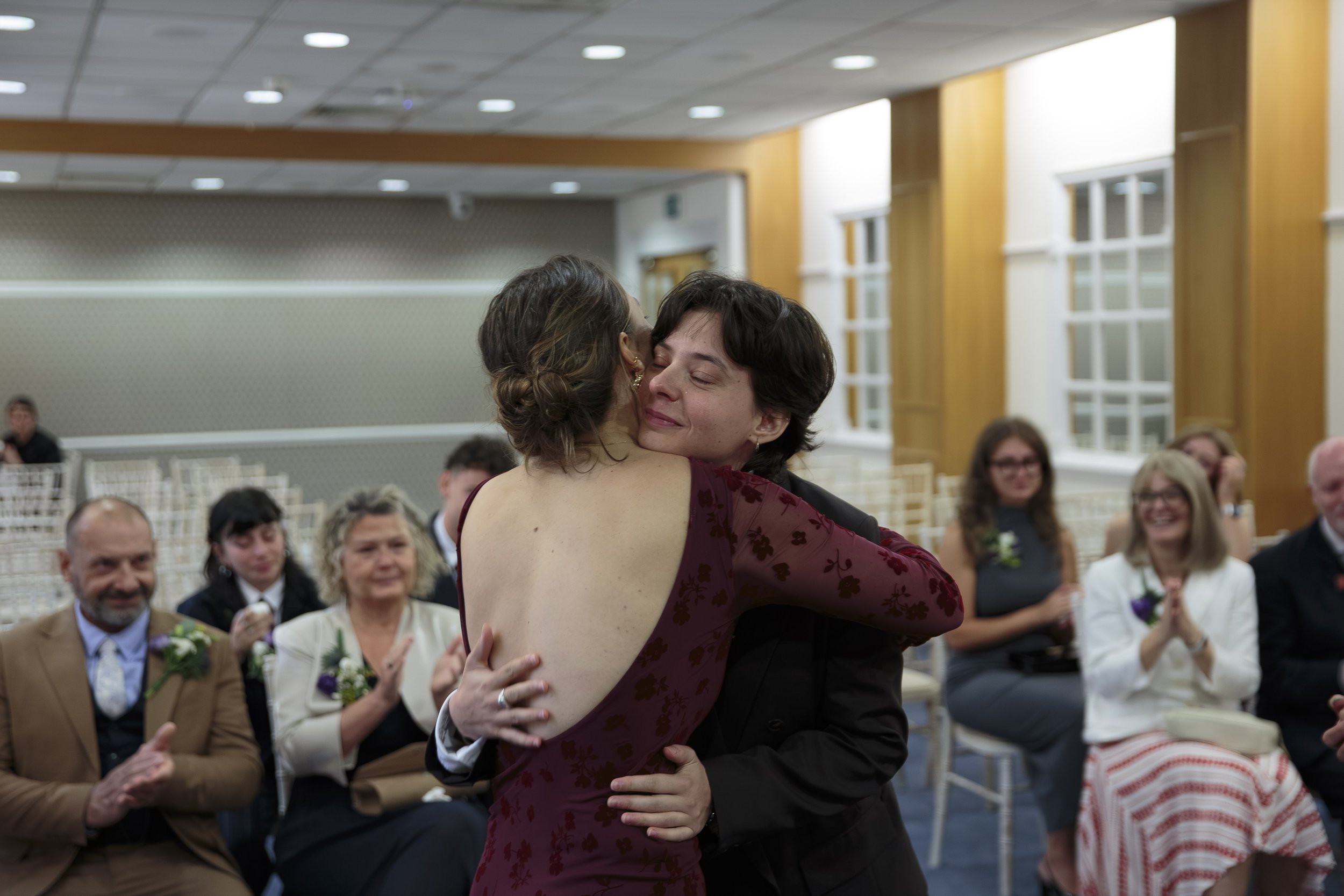 Two women embrace each other in a dance at a formal event, surrounded by seated guests watching and smiling.