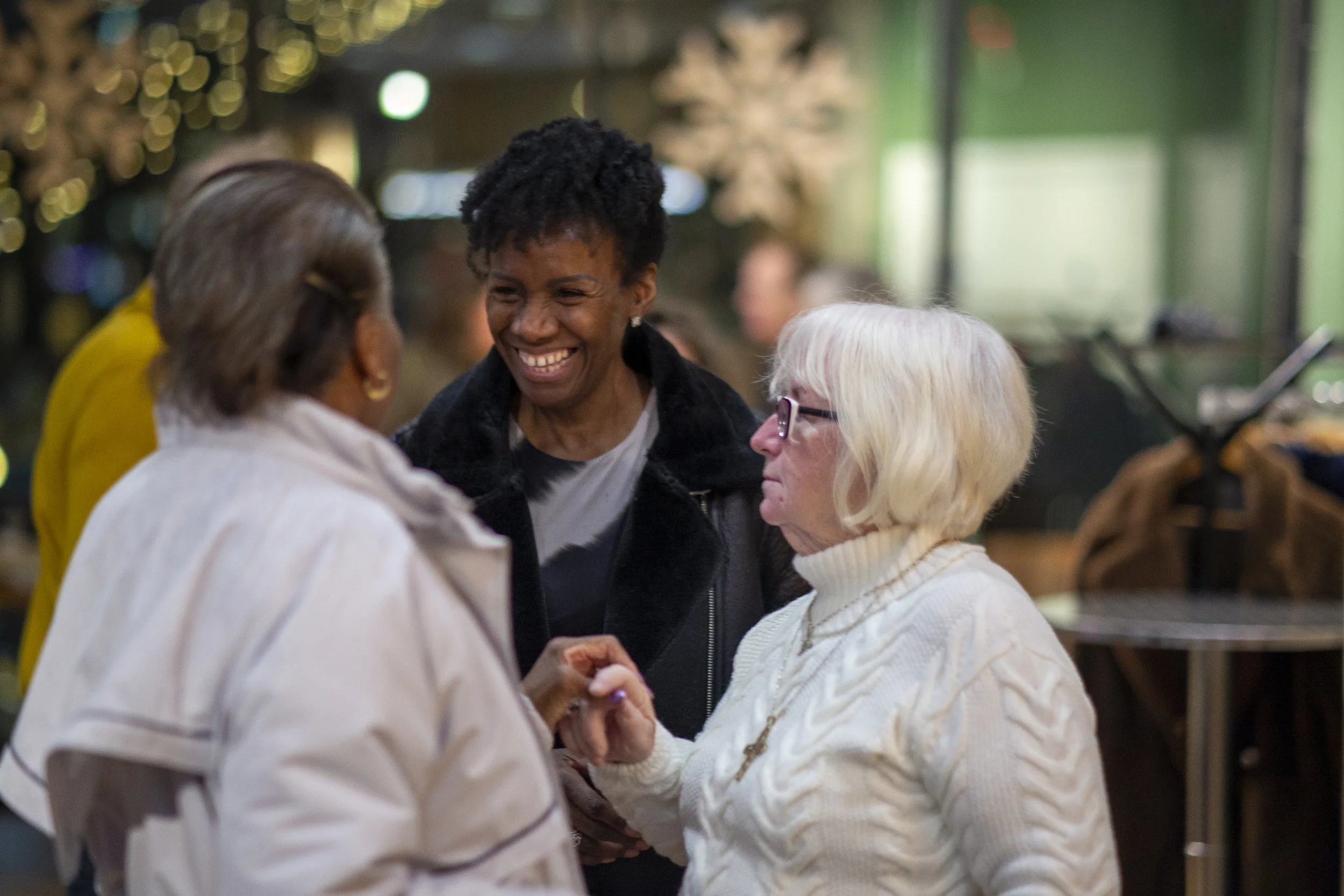 Smiling woman with short curly hair talking to an older woman with white hair at a social gathering indoors, with other people in the background.