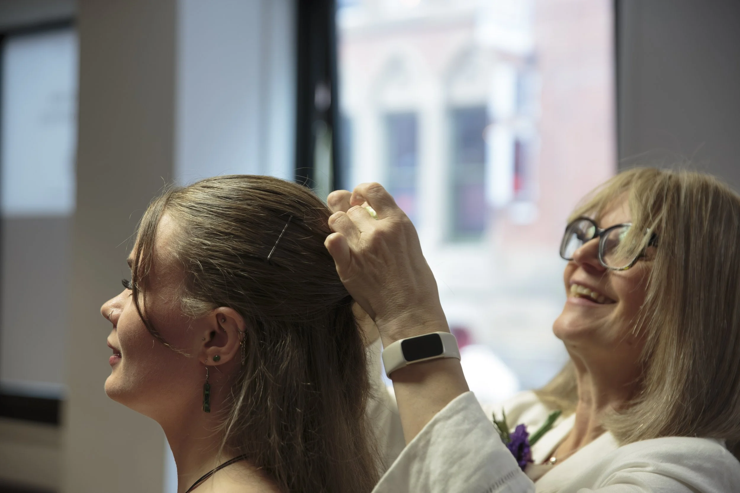 A woman with glasses smiling and a woman with earrings and earrings with her eyes closed, appears to be enjoying a moment together indoors near a window.