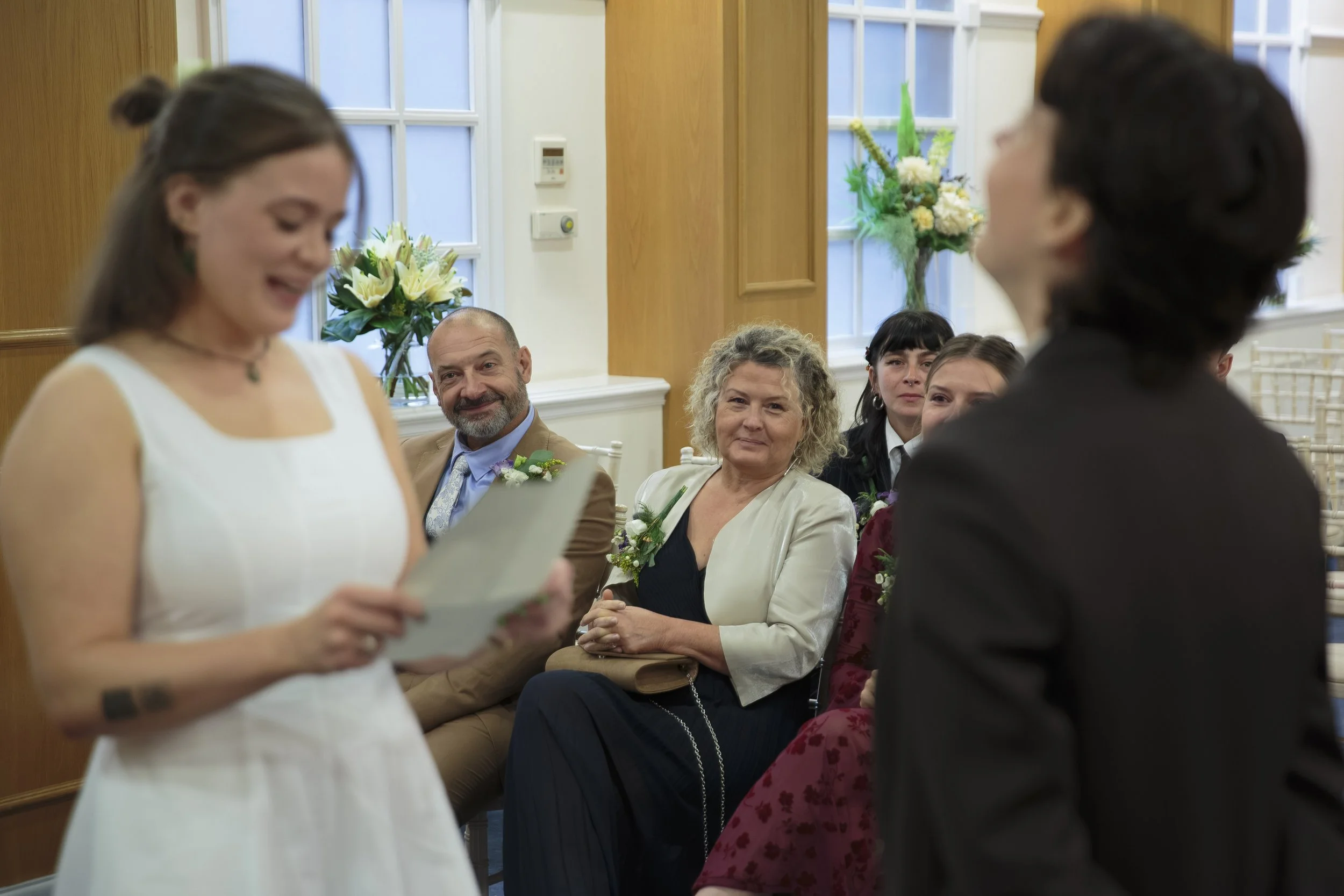 A woman in a white dress appears to be reading from papers during a ceremony, likely a wedding, with a man in a suit and several guests seated and watching attentively. The room is decorated with flowers.
