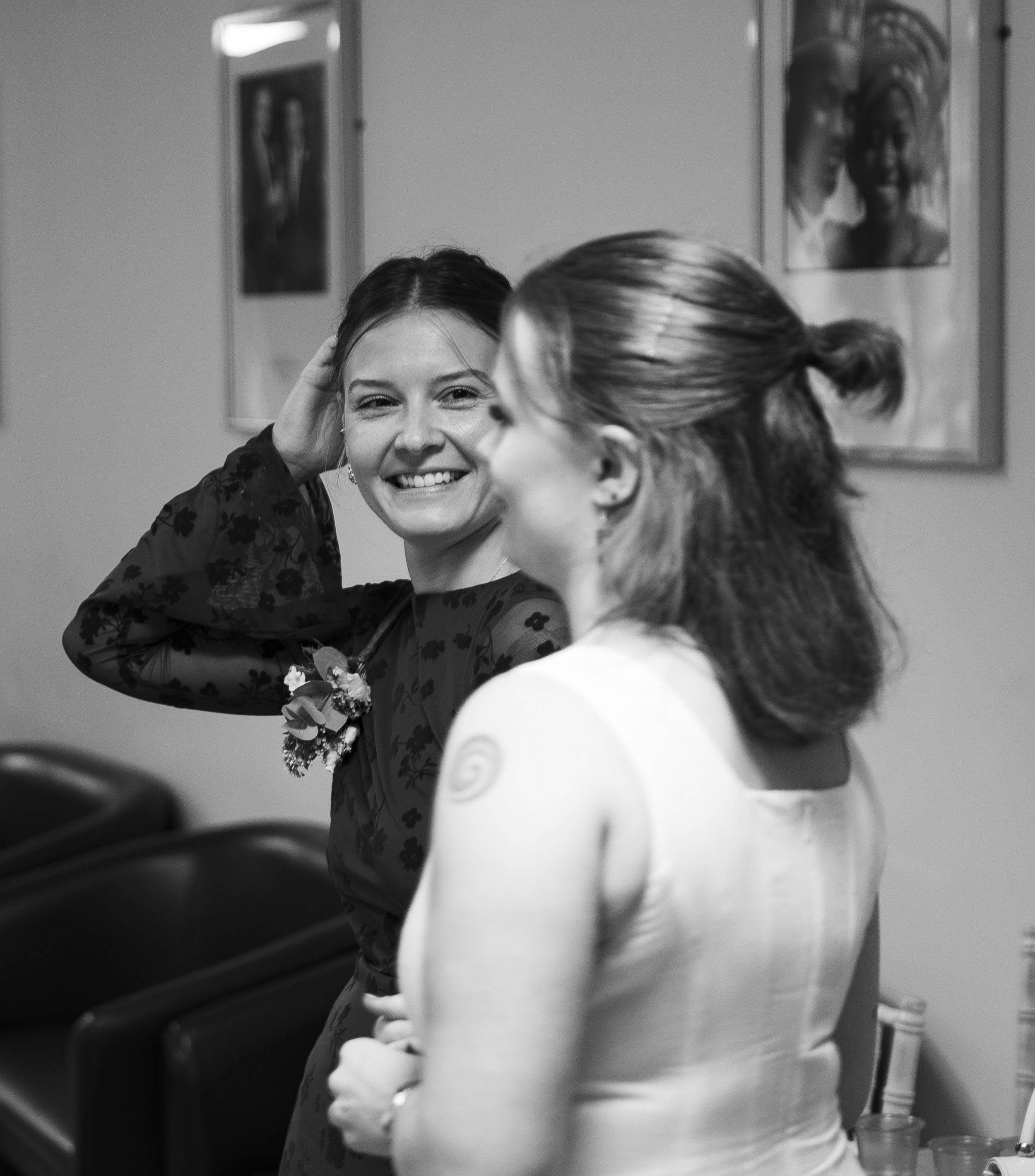 Two women engaged in a conversation, one smiling and the other partially visible, in a setting with framed pictures on the wall, black and white photo.