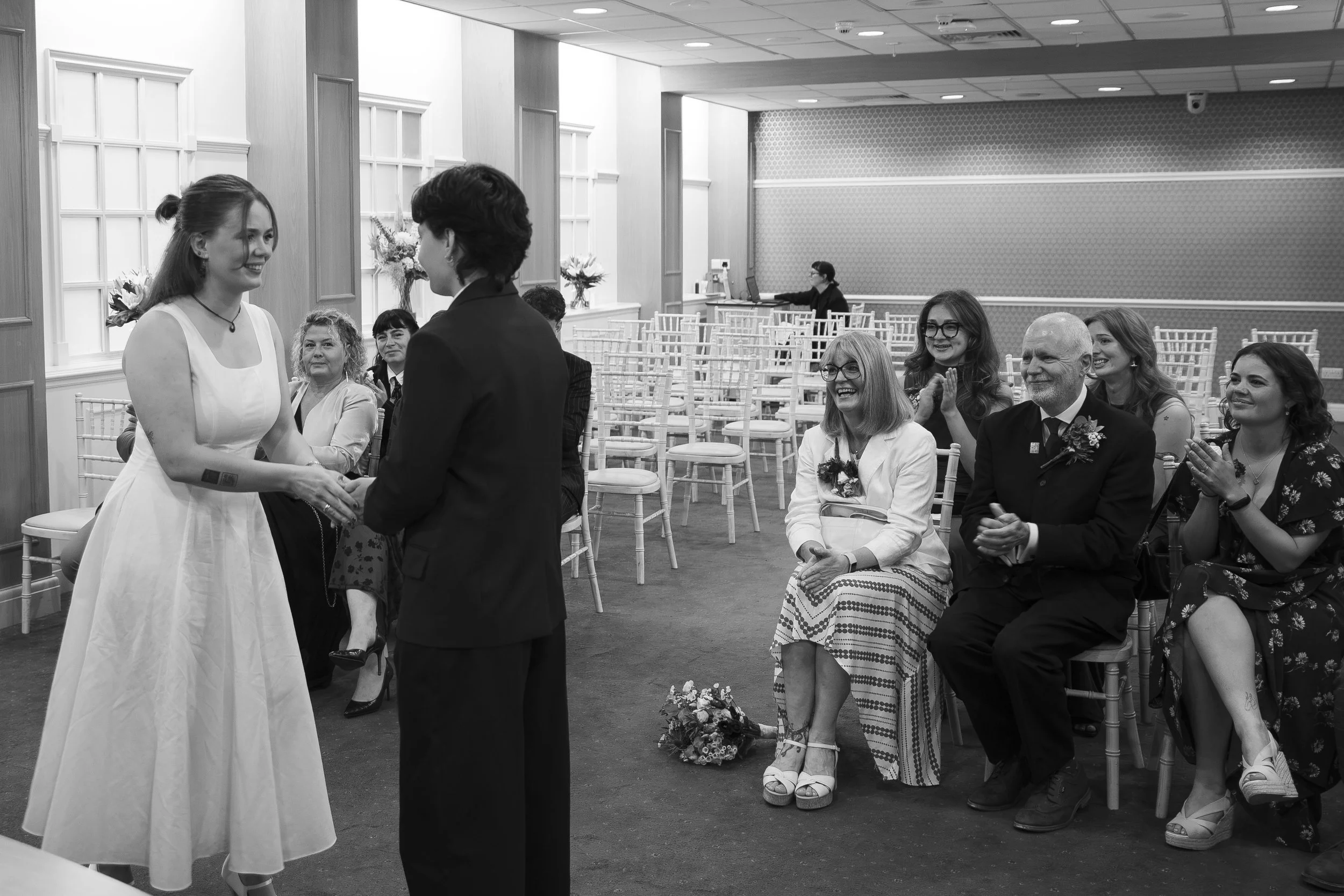A wedding ceremony with a bride and groom shaking hands, guests seated and clapping in a decorated indoor venue.