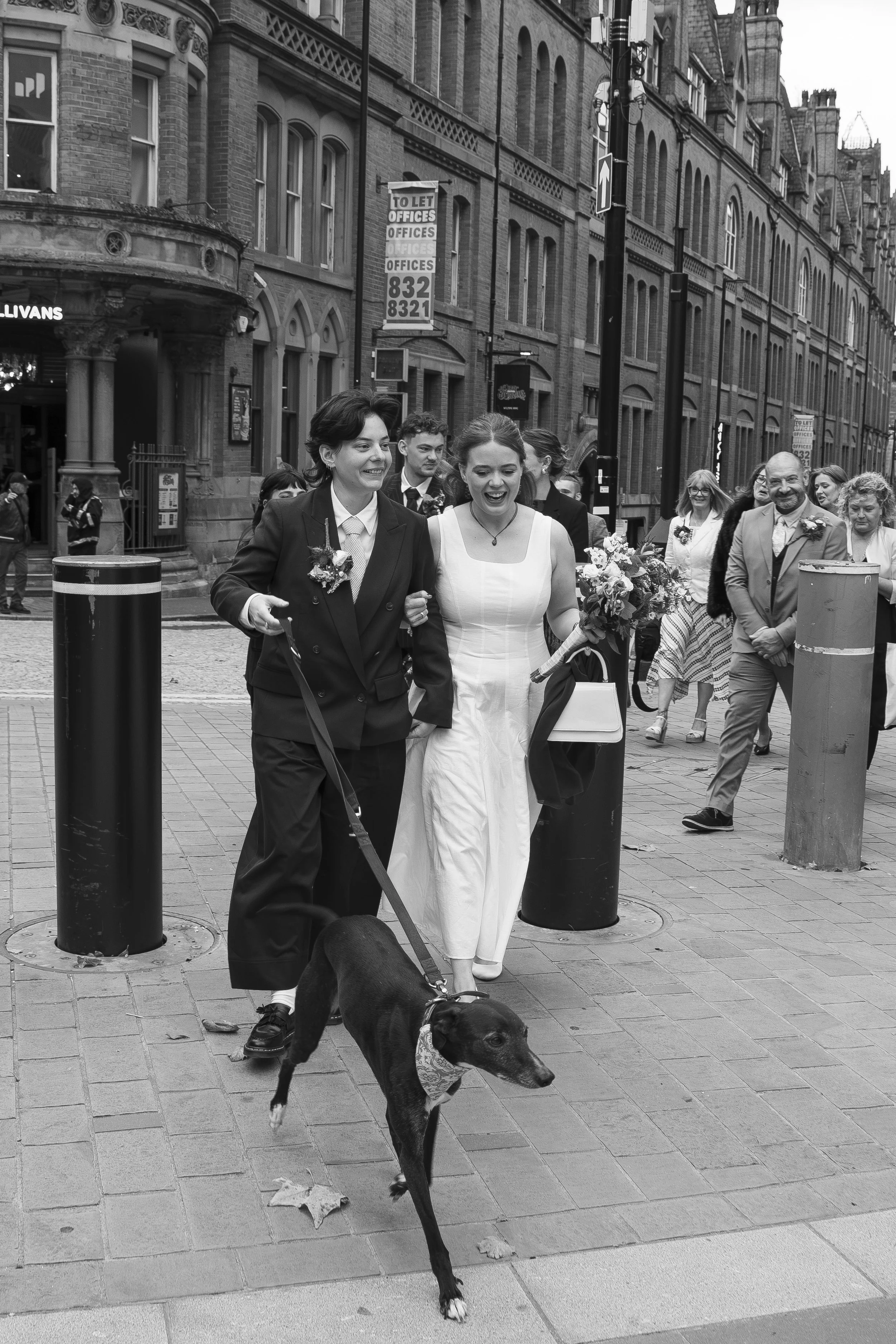 Two women, one in a tuxedo and the other in a white dress, are walking arm in arm on a city street with a group of people following behind them. They are smiling and holding flowers. A black dog is walking ahead of them. The setting is urban with bri