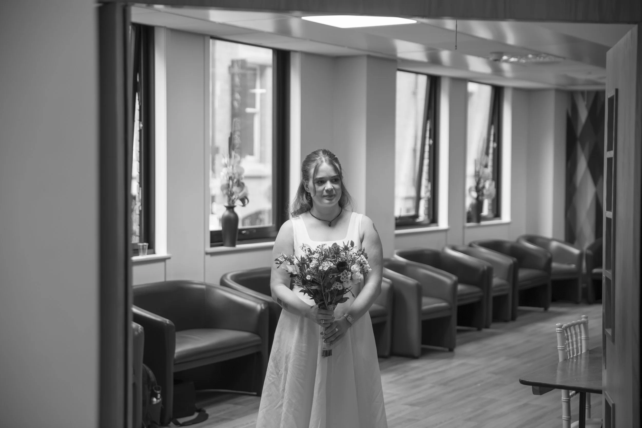 Black and white photo of a woman in a dress holding a bouquet of flowers, standing in a room with large windows and leather chairs.