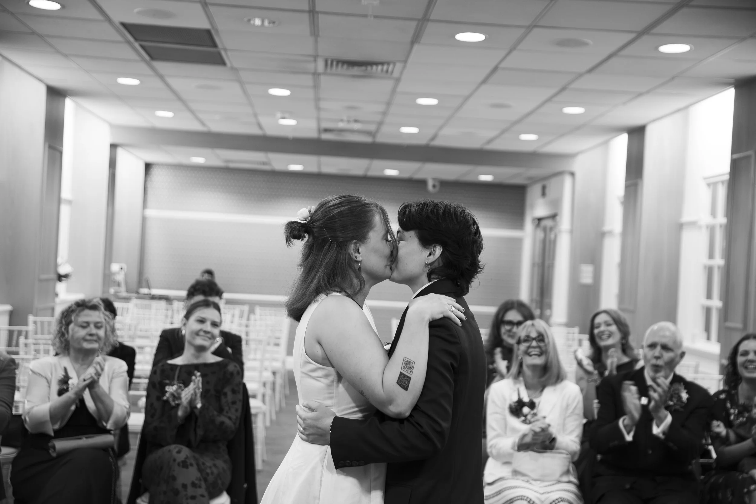 Two women sharing a kiss at what appears to be a wedding ceremony with seated guests clapping and smiling in the background.